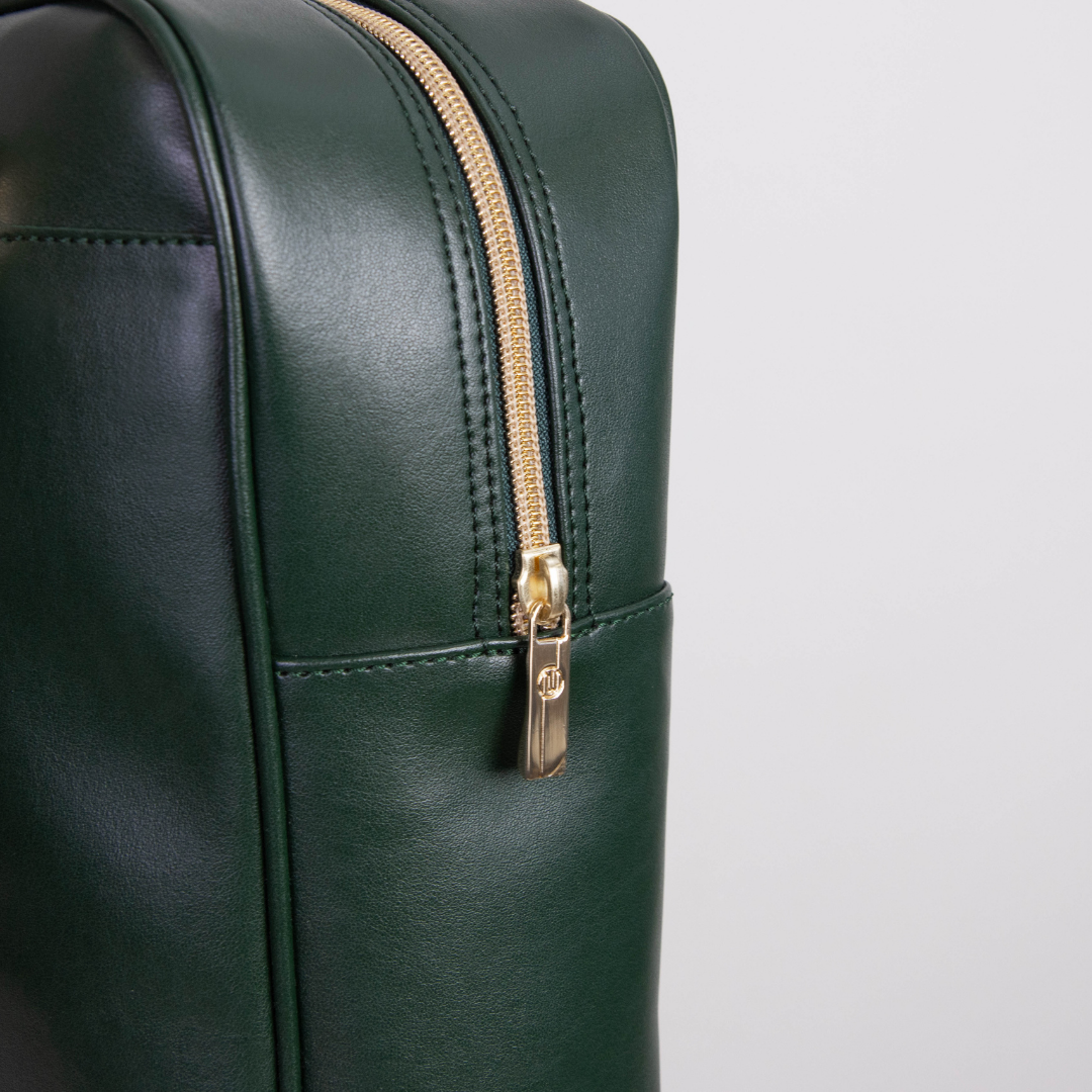 Dunkelgrüne Ledertasche mit goldenem Reißverschluss und dezentem Logo auf dem Zipper, fotografiert vor neutralem Hintergrund.