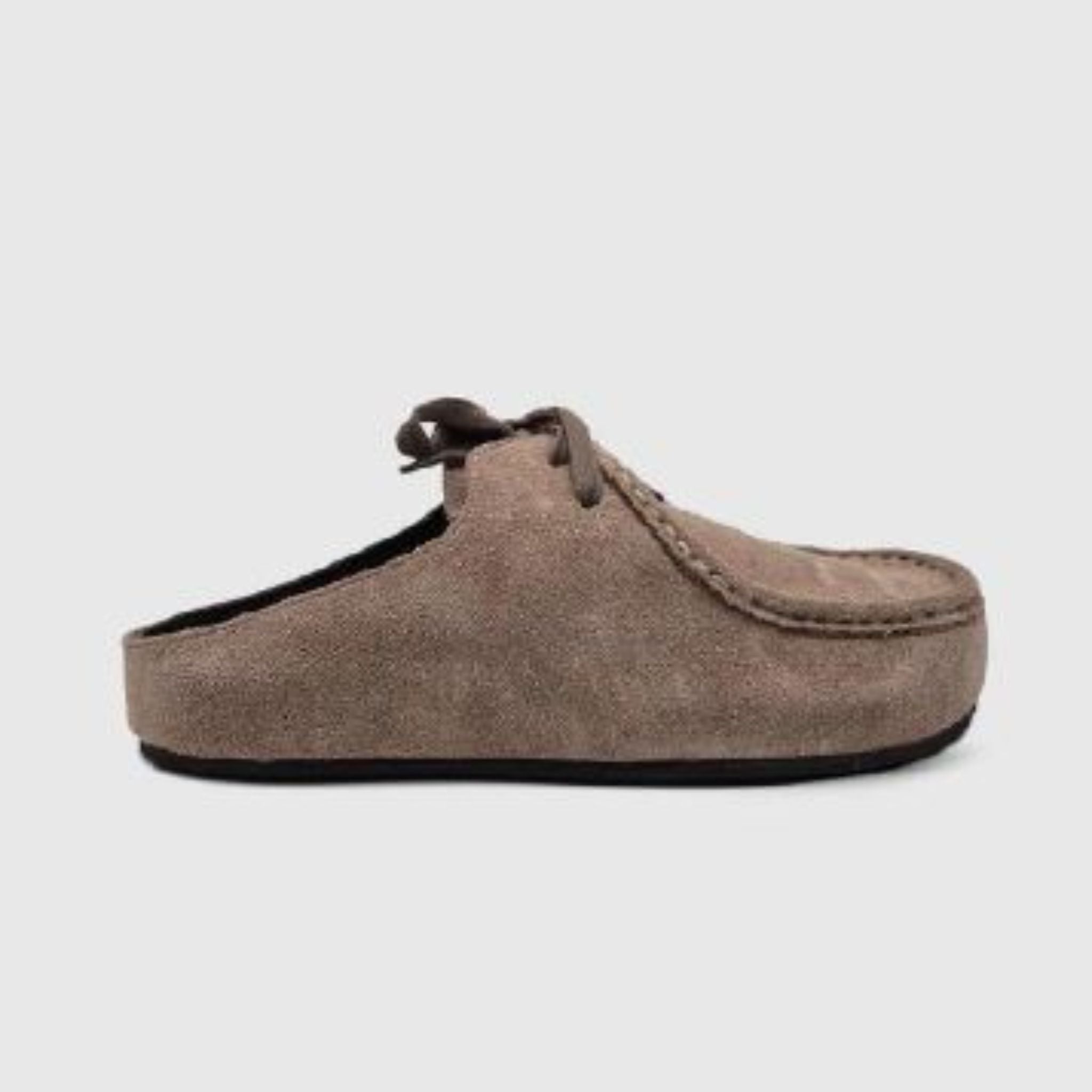 Ein brauner Slipper aus Wildleder mit geschlossener Zehenpartie, sichtbaren Nähten, dekorativen Schnürsenkeln und einer flachen schwarzen Sohle, im Profil auf hellgrauem Hintergrund.