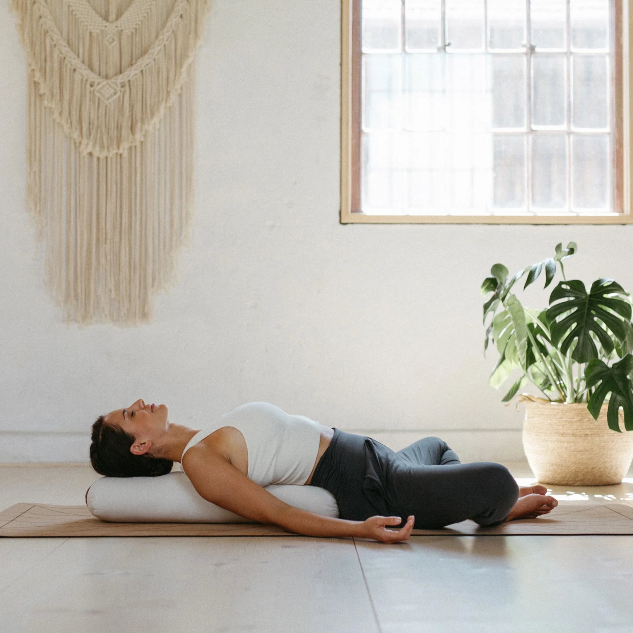 Person liegt auf einem Yoga-Bolster in einem hellen Raum, mit einer Zimmerpflanze und einer Makramee-Wanddekoration.
