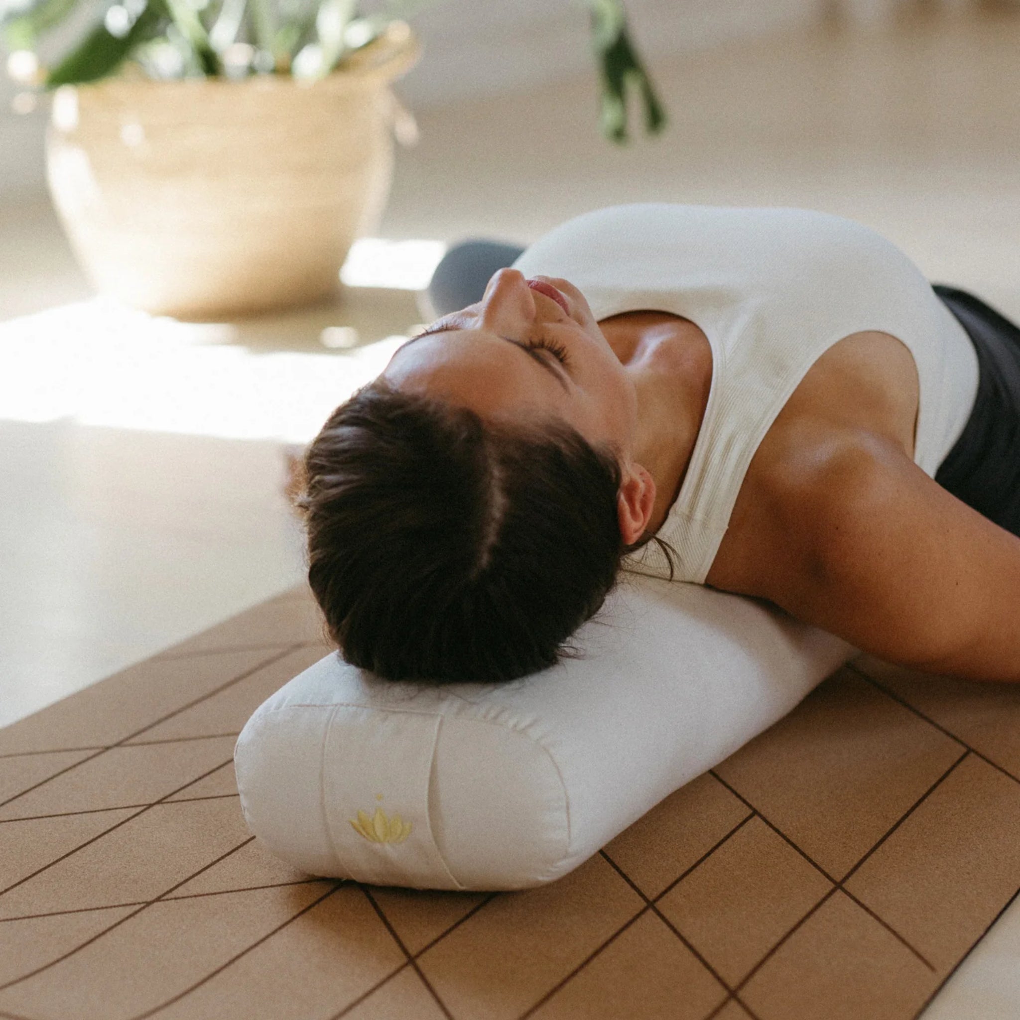 Frau liegt entspannt auf einem Yoga-Bolster auf einer Matte, im Hintergrund eine Pflanze.