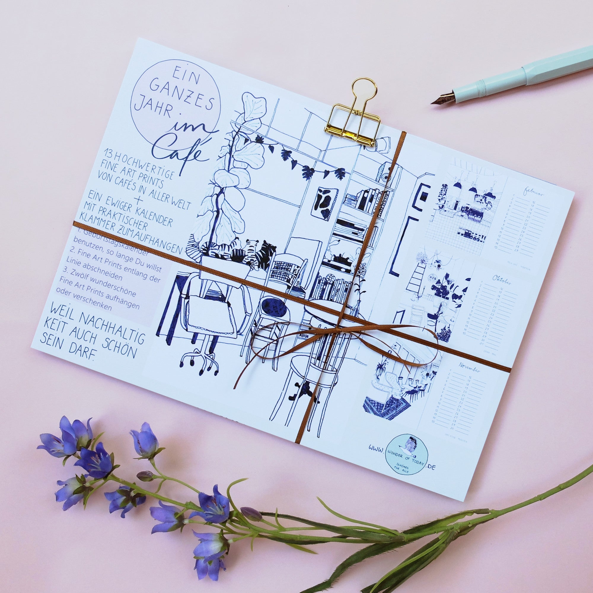 Ein Kalenderblatt mit handgezeichneten Illustrationen eines gemütlichen Café-Interieurs, deutschem Text und einer goldenen Klammer, platziert auf einer rosa Fläche mit blauen Blumen und einem blaugrünen Stift in der Nähe.