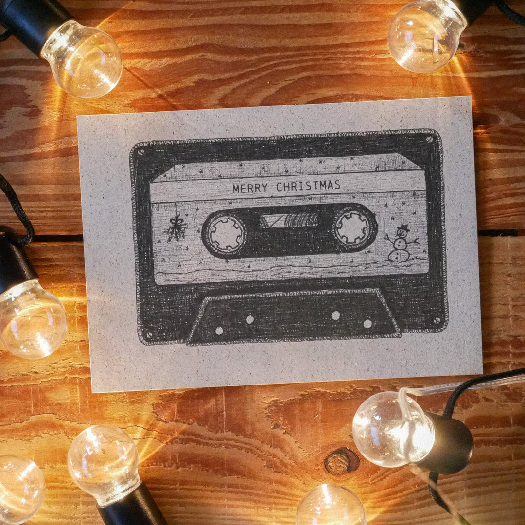 Karte mit Kassettendesign und "Merry Christmas"-Text, umgeben von Lichterketten auf einem Holztisch.