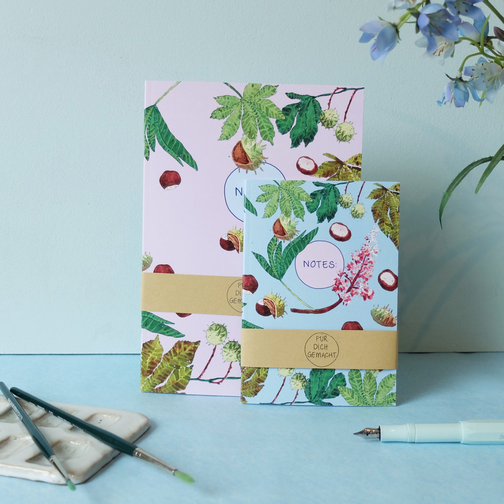 Zwei bunte Notizbücher mit Kastanien- und Blättermotiven, daneben ein Pinsel, eine Palette und ein Stift auf blauem Untergrund.