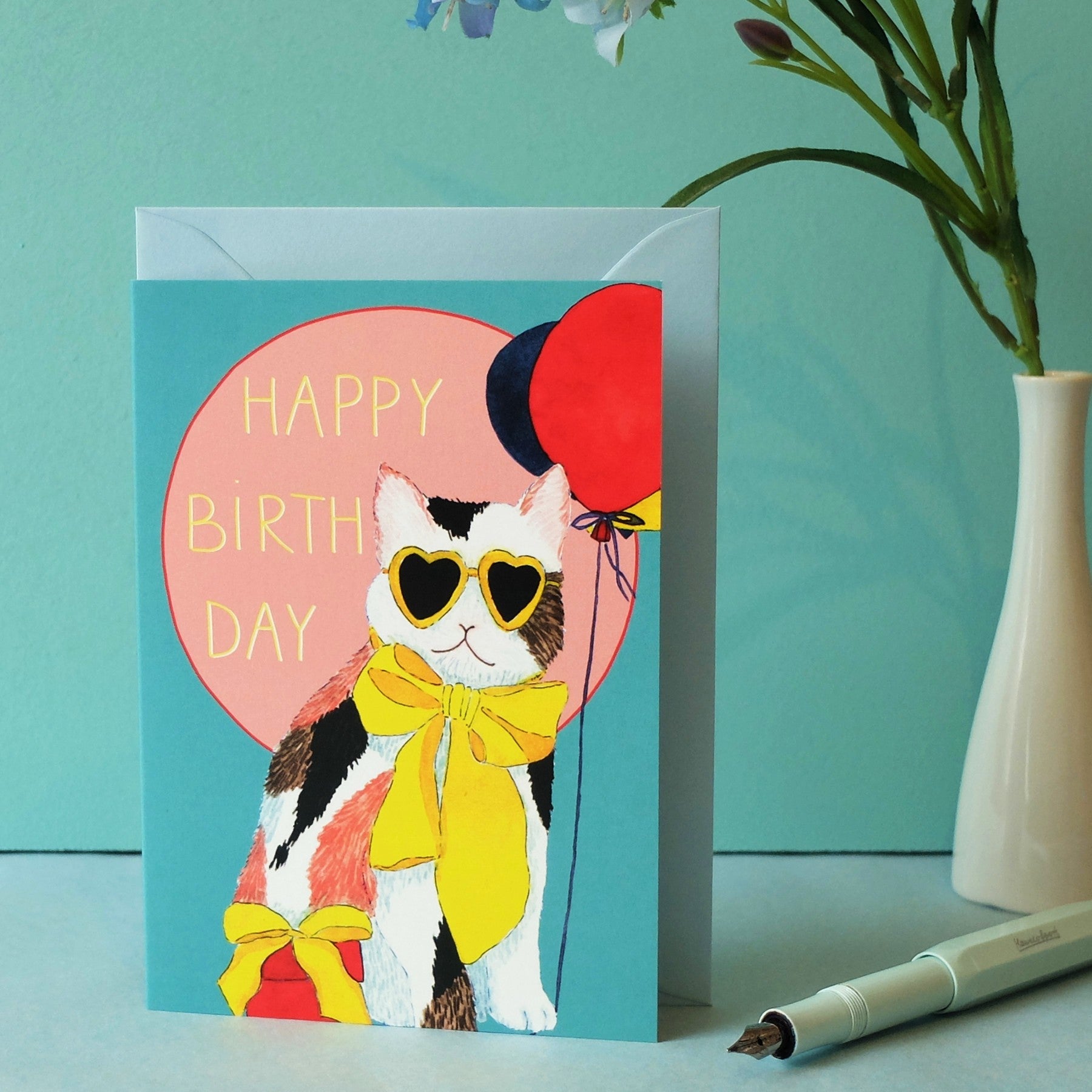 Grußkarte mit einer illustrierten Katze, die eine herzförmige Sonnenbrille und eine gelbe Schleife trägt und zwei Luftballons hält. Der Text auf der Karte lautet HAPPY BIRTHDAY. In der Nähe stehen eine Vase mit Blumen und ein Stift.