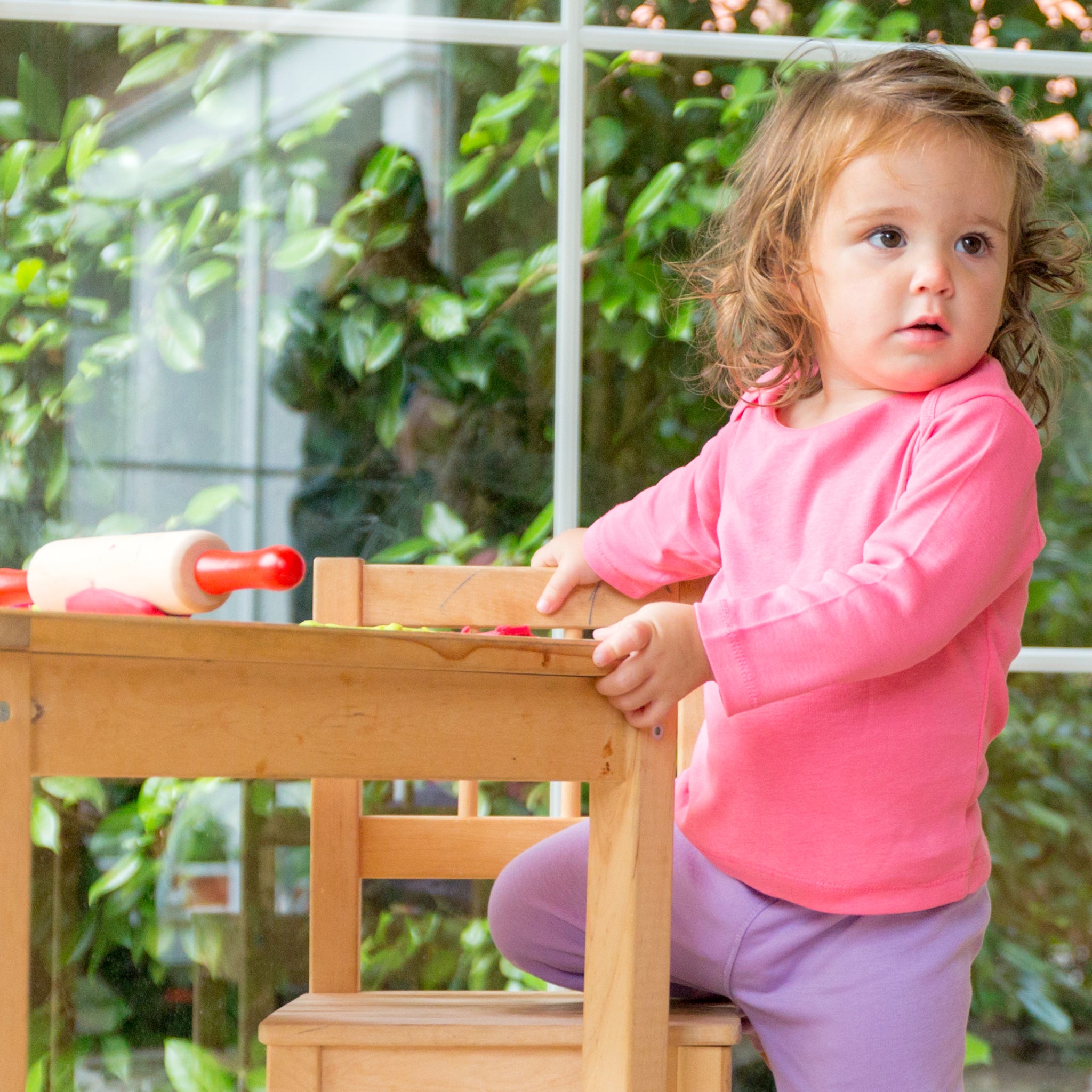 Ein kleines Kind in einem rosa Oberteil steht neben einem Holztisch mit Nudelholz und Spielzeug vor einem Fenster.