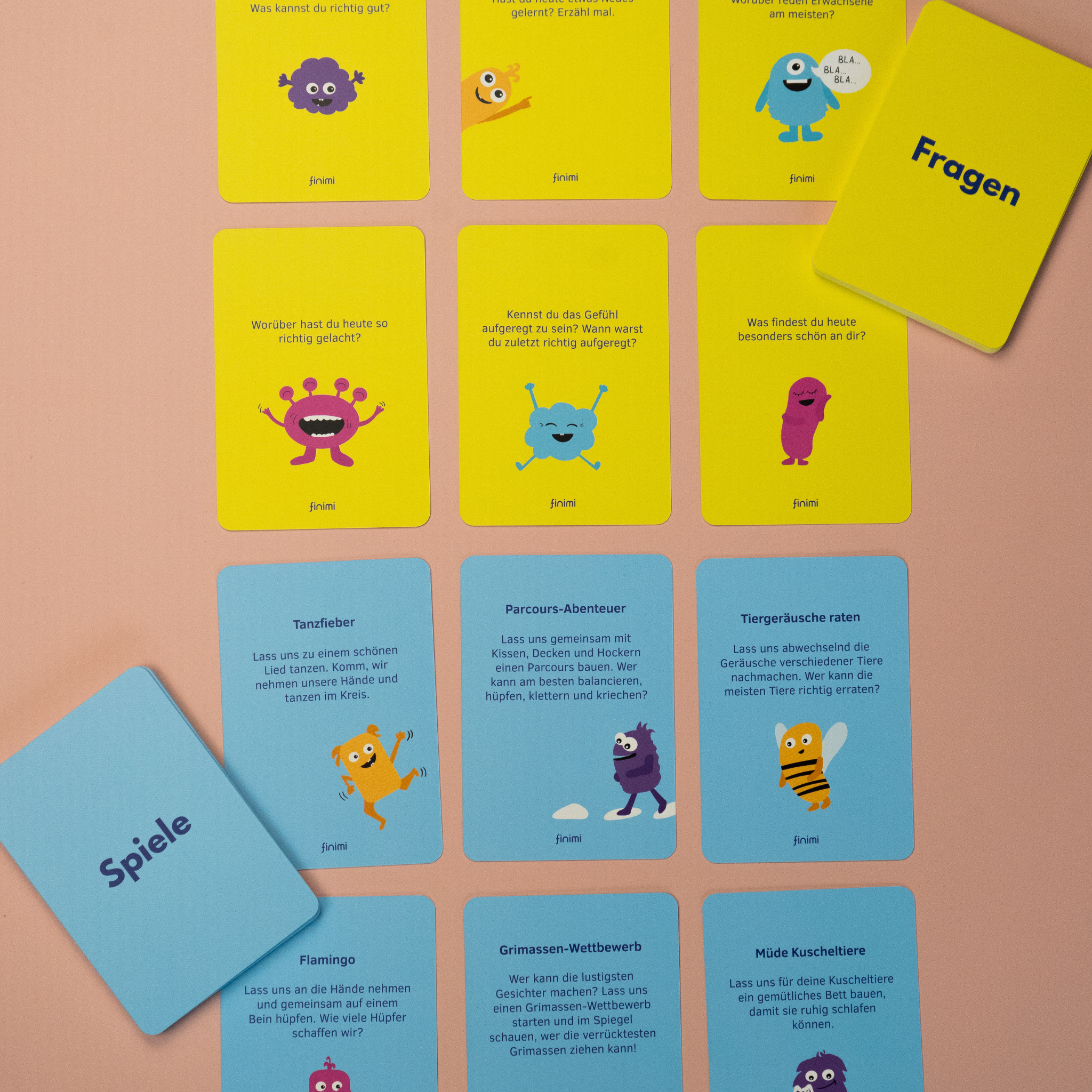 Eine Sammlung bunter Spielkarten in deutscher Sprache ist auf einer rosa Fläche ausgebreitet. Die Karten zeigen verspielte Monster-Illustrationen und sind mit "Fragen" und "Spiele" beschriftet.