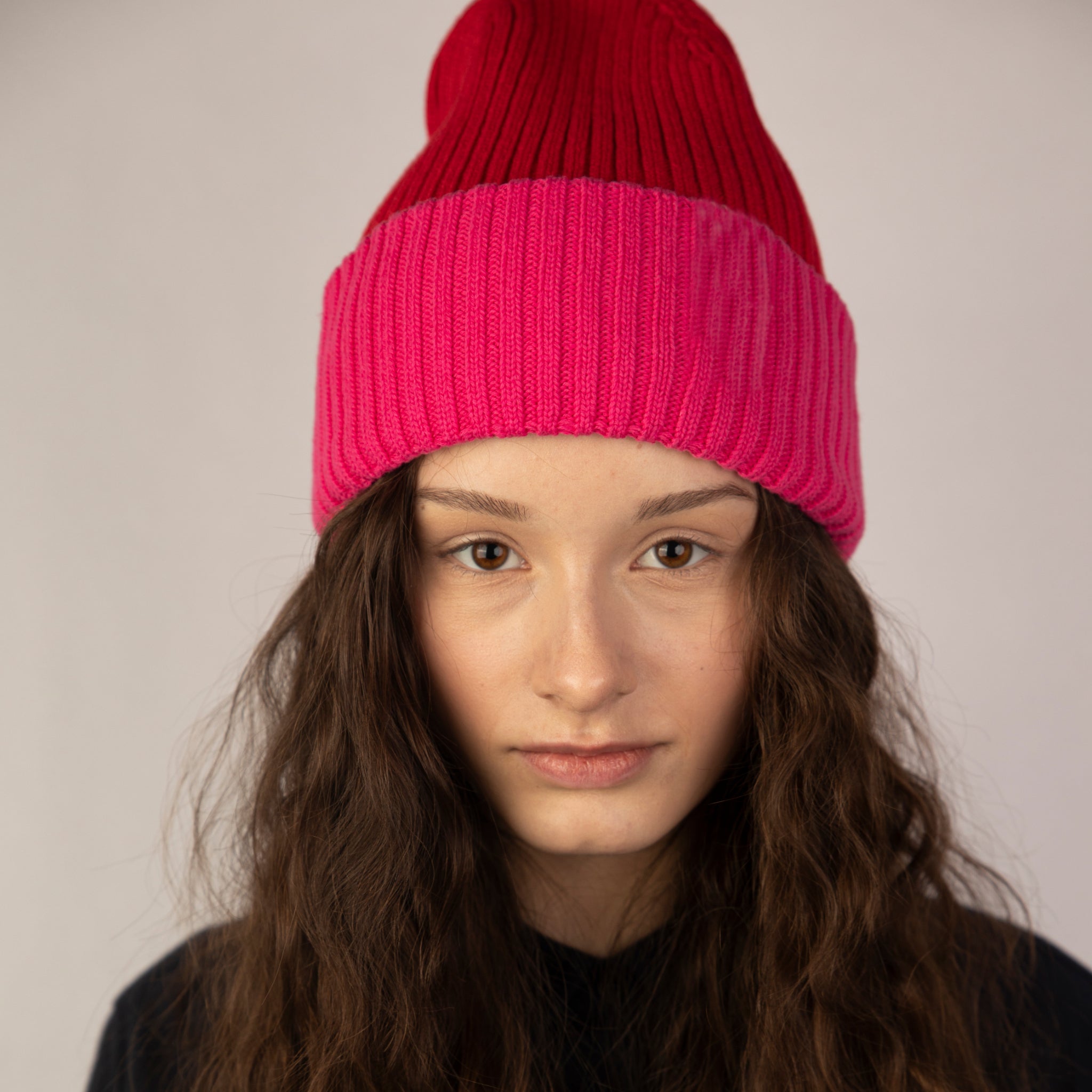 Eine Person mit langen, gewellten braunen Haaren trägt eine rot-rosafarbene Strickmütze und ein schwarzes Hemd und schaut mit neutralem Gesichtsausdruck direkt in die Kamera vor einem schlichten hellen Hintergrund.