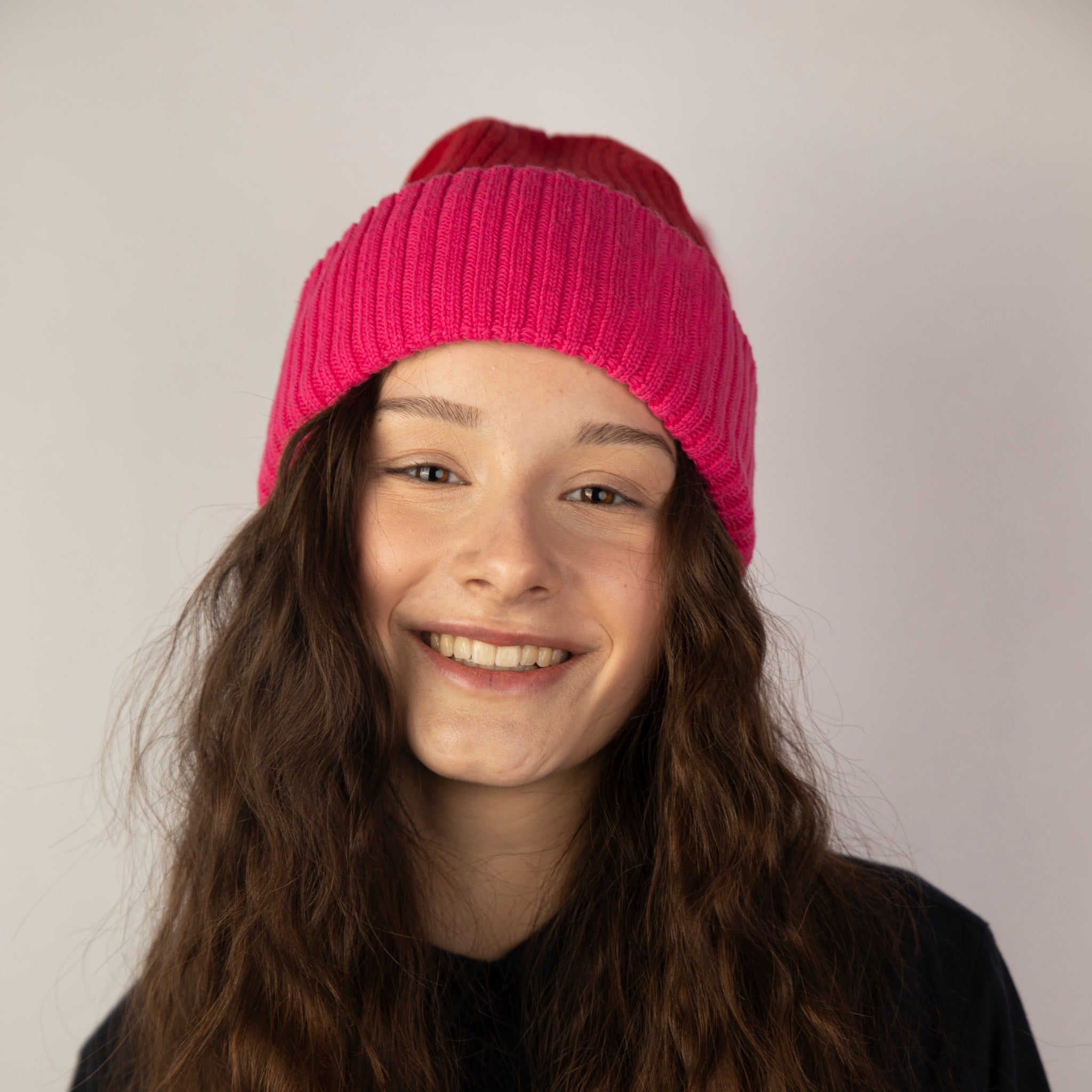 Eine junge Person mit langen, gewellten braunen Haaren lächelt in die Kamera, während sie eine hellrosa und rote Strickmütze und ein schwarzes Hemd vor einem schlichten, hellen Hintergrund trägt.