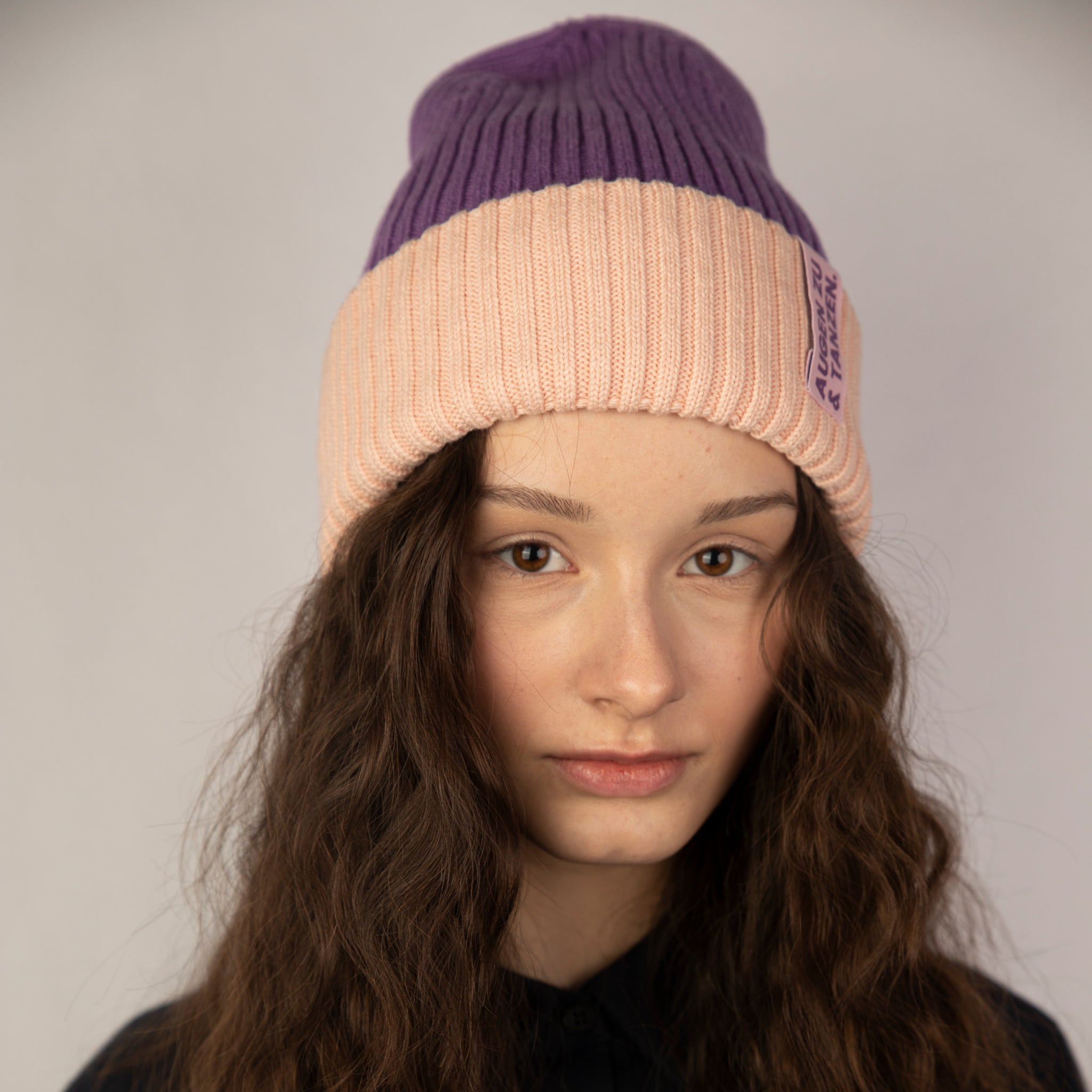 Junge Frau mit langen, gewellten braunen Haaren, die eine lila und pfirsichfarbene Strickmütze trägt und mit neutralem Gesichtsausdruck direkt in die Kamera schaut, vor einem schlichten Hintergrund.