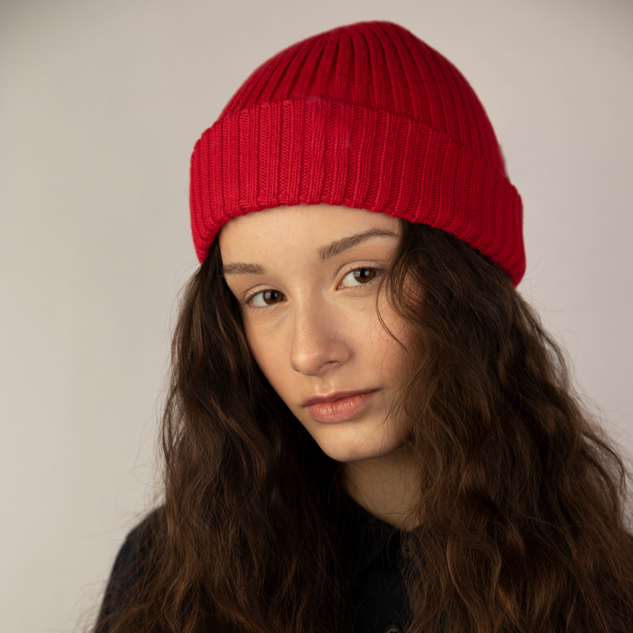 Eine junge Frau mit langen, gewellten braunen Haaren trägt eine leuchtend rote Strickmütze und ein schwarzes Oberteil und blickt ruhig in die Kamera vor einem schlichten, hellen Hintergrund.