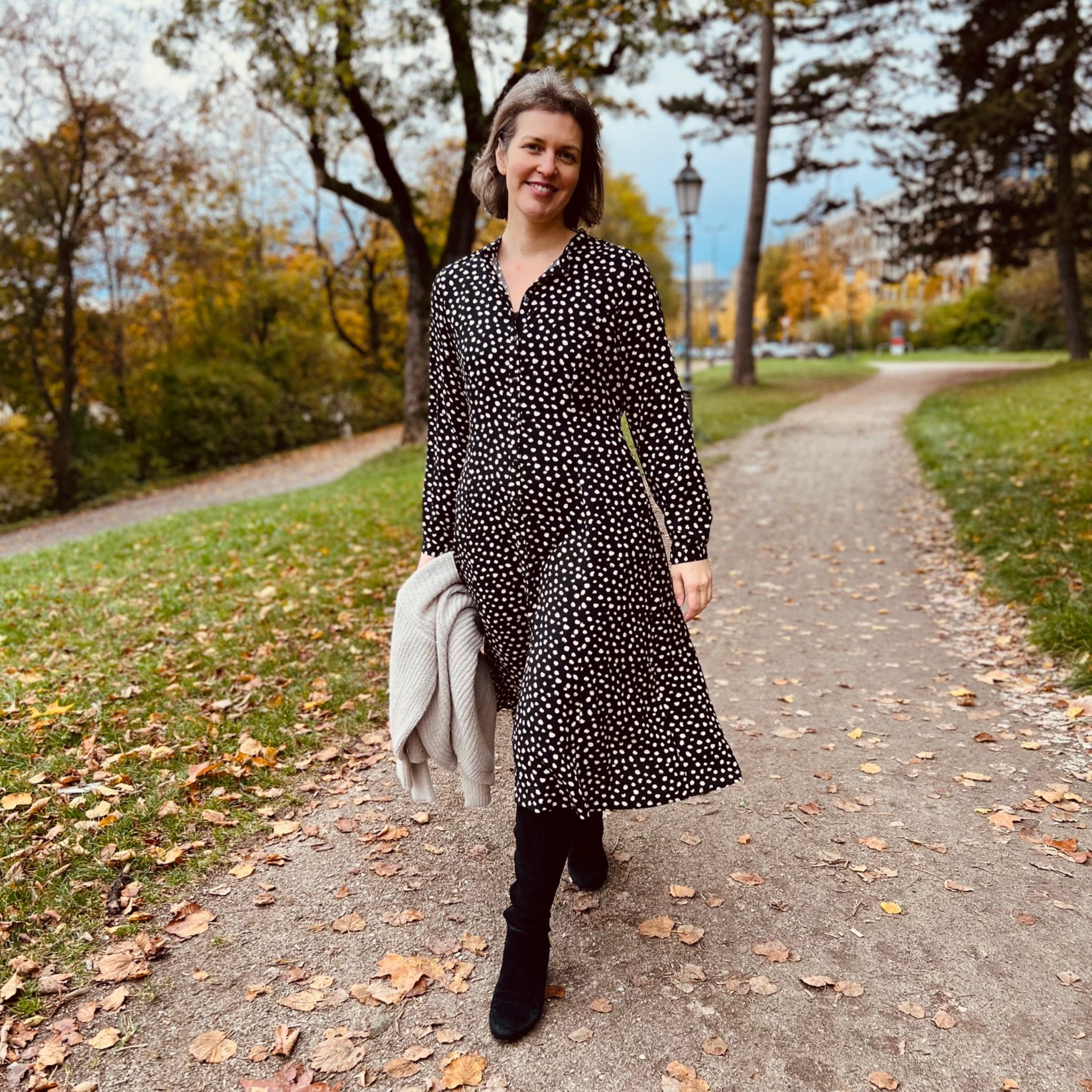 Frau in gepunktetem Kleid spaziert auf einem Herbstweg, umgeben von bunten Bäumen und Laub.