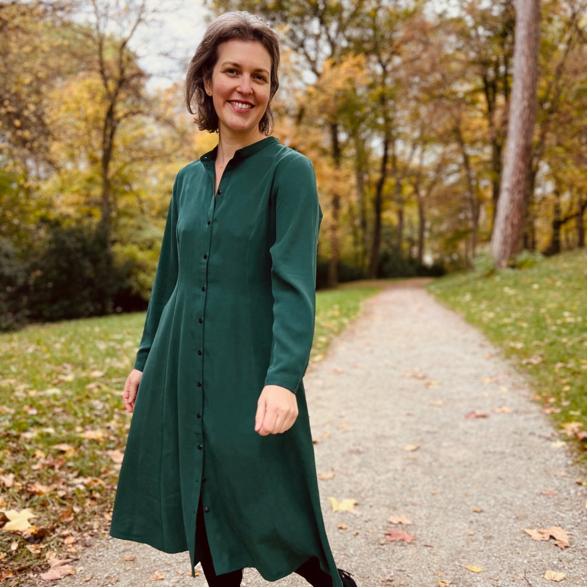 Frau in grünem Kleid geht lächelnd auf einem Weg im herbstlichen Park spazieren.