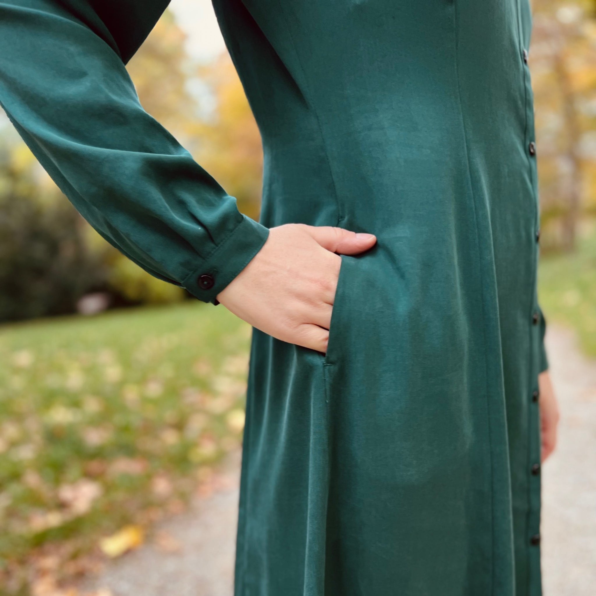 Nahaufnahme einer Person, die ein grünes langes Kleid mit Knöpfen trägt und eine Hand in der Tasche hält.