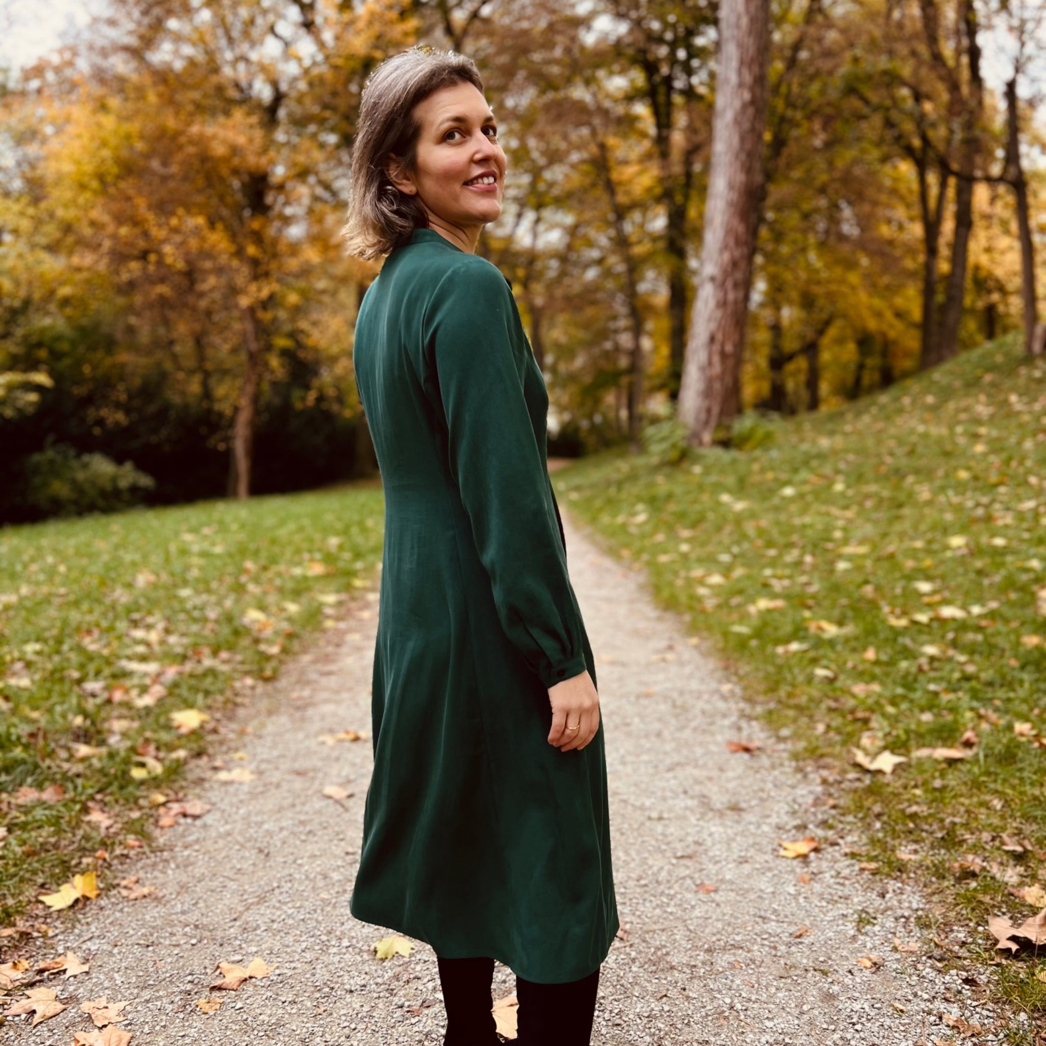 Frau in grünem Kleid steht auf einem Herbstweg im Wald und blickt über die Schulter zurück.