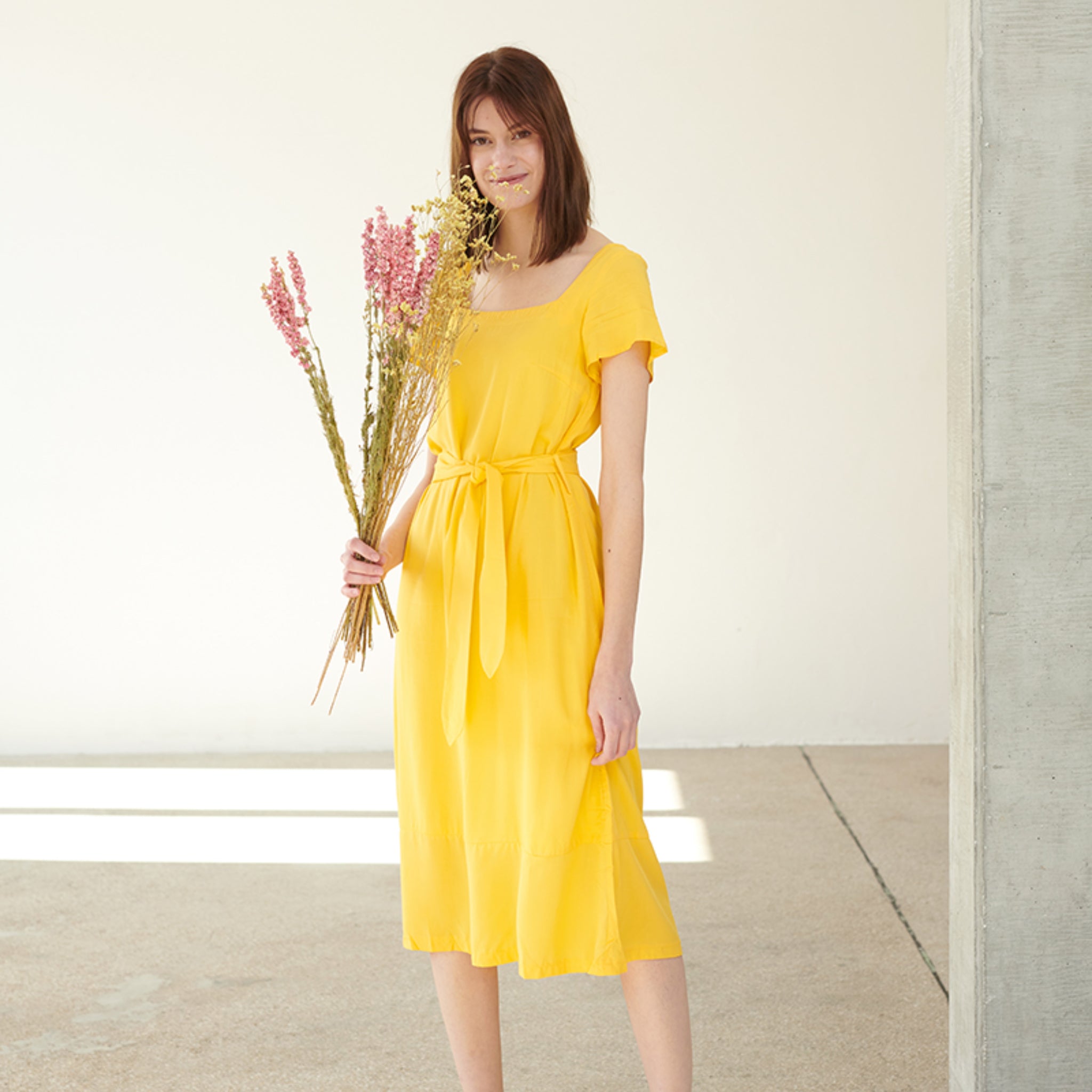 Frau in gelbem Kleid hält einen Strauß aus Trockenblumen in einem minimalistischen Raum.