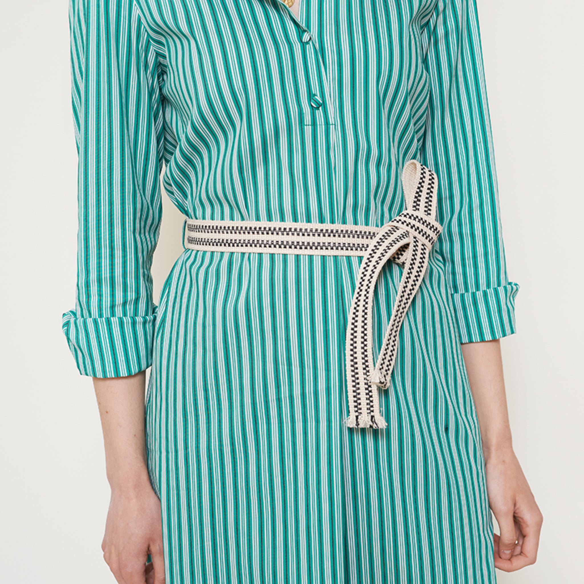 Person in gestreiftem, grünem Kleid mit weißen und schwarzen Gürtel, die Arme seitlich herabhängend.