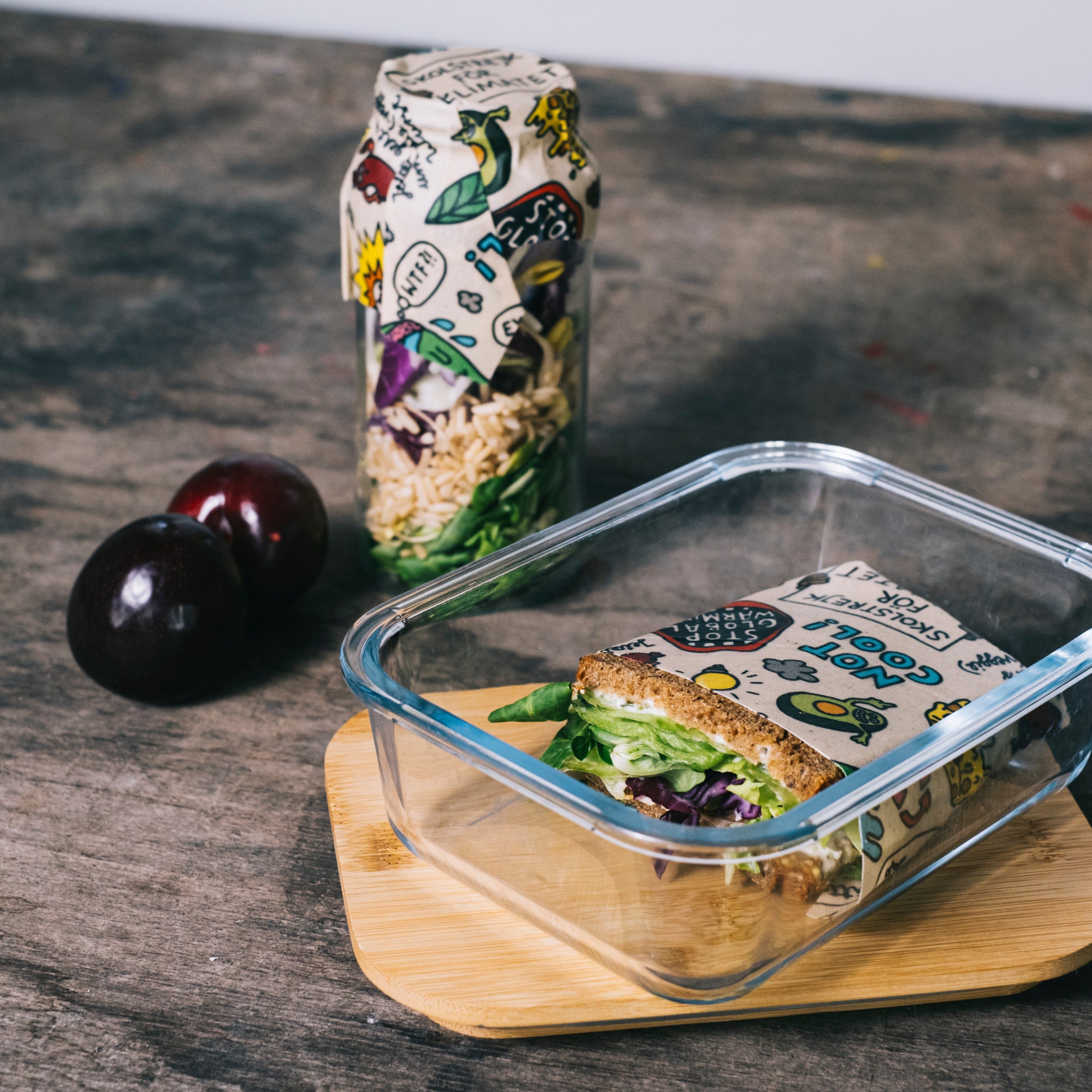 Ein Sandwich und Obst auf Holzbrett neben einer dekorativen Glasflasche mit Salat auf einer rustikalen Tischoberfläche.