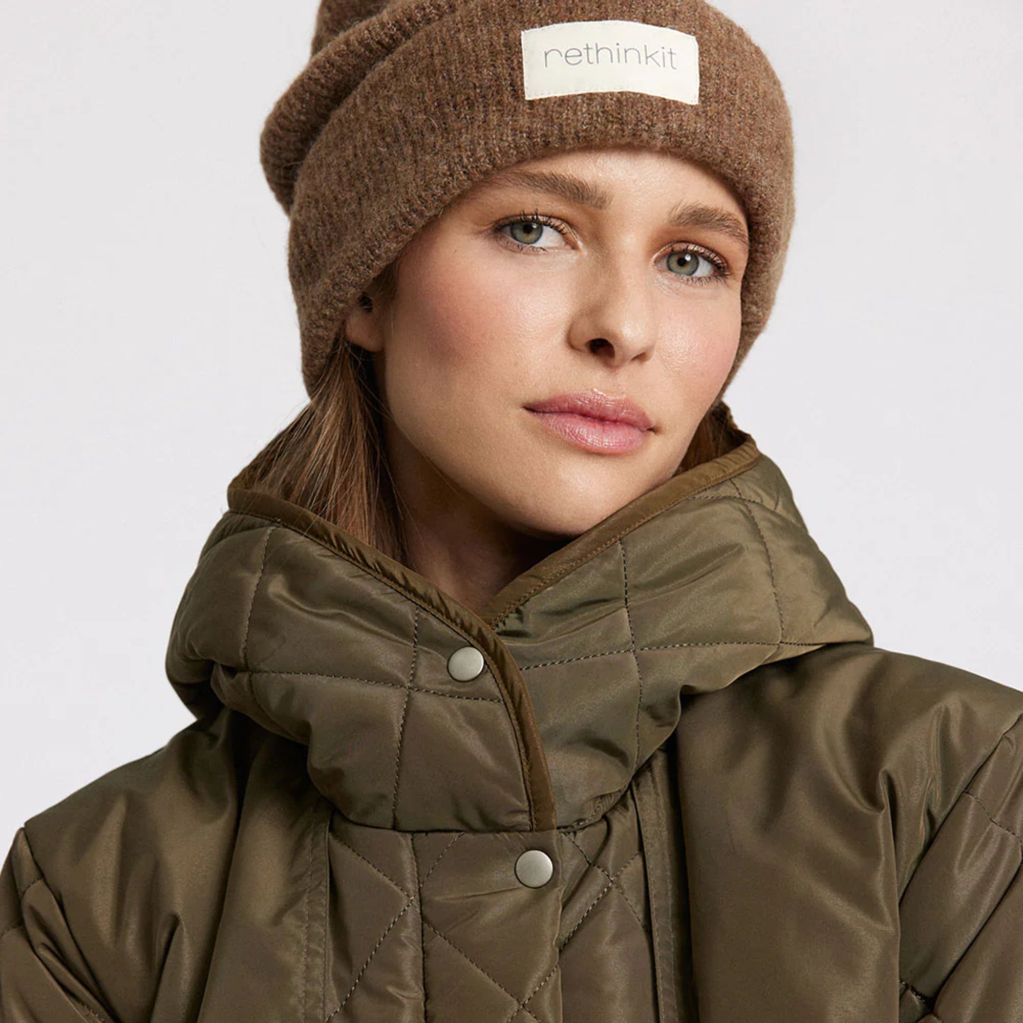 Eine Frau, die eine braune Strickmütze mit einem Rethinkit-Etikett und eine olivgrüne Steppjacke trägt, blickt vor einem schlichten hellen Hintergrund in die Kamera.