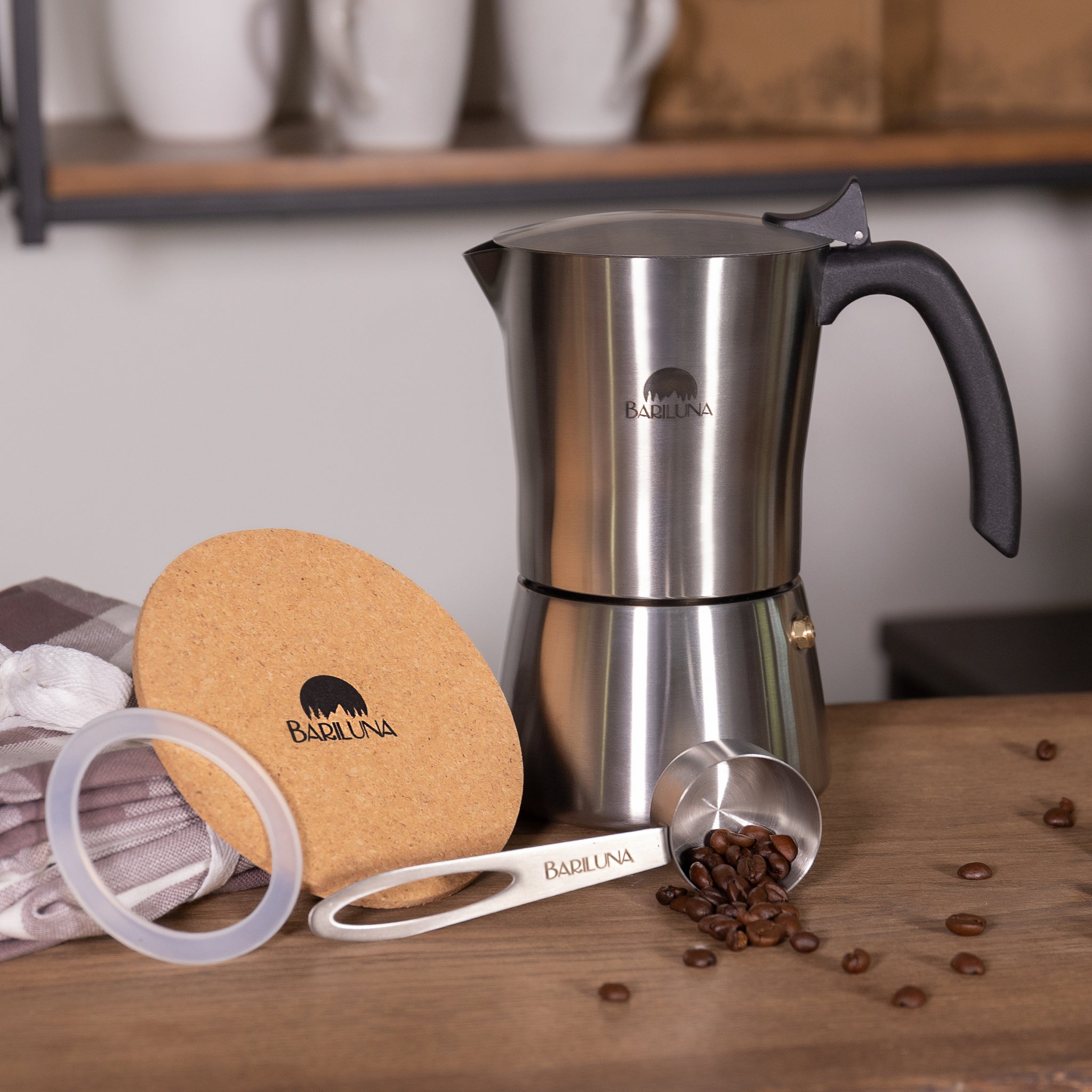 Edelstahl-Kaffeemaschine mit schwarzem Griff, Korkuntersetzer, Maßlöffel mit Kaffeebohnen und Geschirrtuch auf Holzoberfläche.