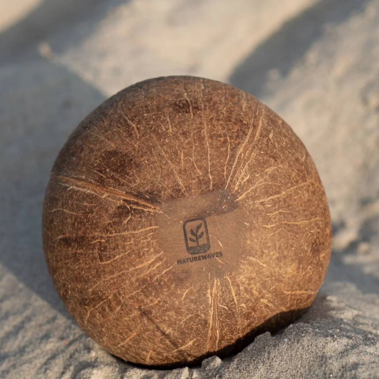 Kreisförmiger, kokosnussartiger Ball mit „Naturewaves“-Logo liegt im Sand.