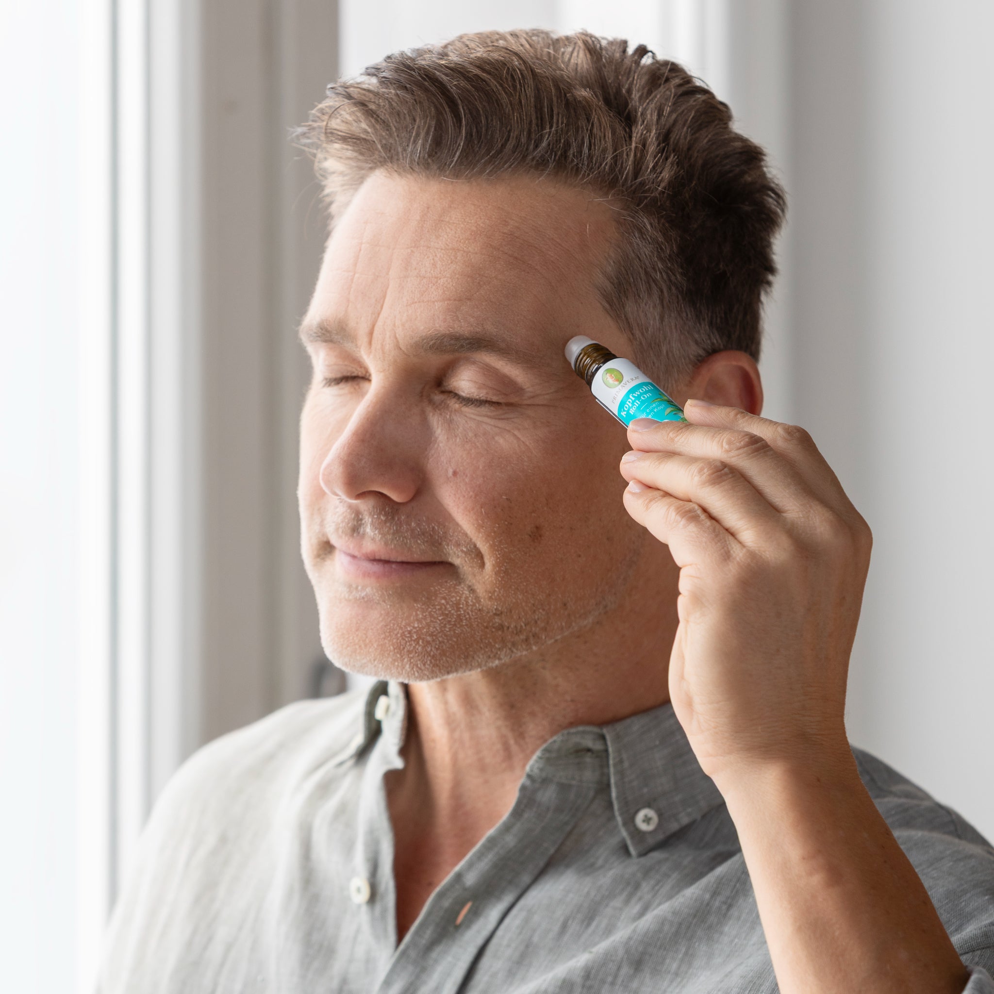 Ein Mann mit kurzen braunen Haaren und geschlossenen Augen hält sich eine kleine Rollerflasche an die Schläfe. Er steht an einem hellen Fenster und trägt ein hellgraues Hemd.