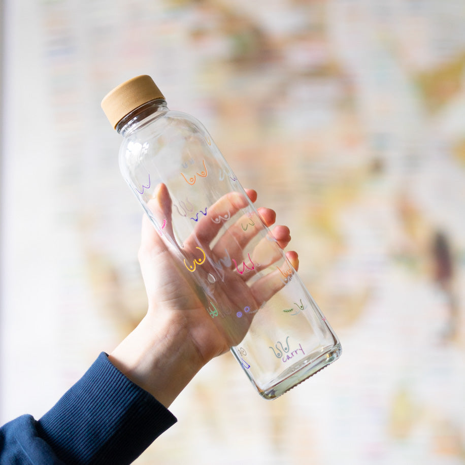 Hand hält eine Glasflasche mit einem Holzdeckel, dekoriert mit bunten Linienmotiven, vor einem verschwommenen Hintergrund.