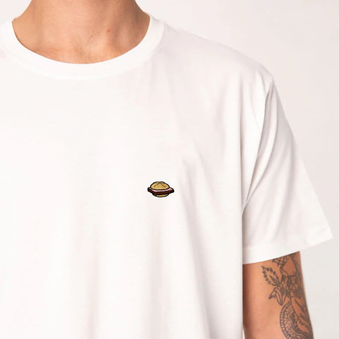 Eine Person, die ein schlichtes weißes T-Shirt mit einem kleinen, aufgestickten Planetenmuster auf der linken Brust trägt. Nur die untere Hälfte des Gesichts, der obere Teil des Oberkörpers und der tätowierte rechte Arm sind sichtbar.