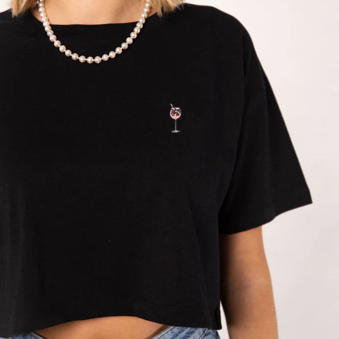 Eine Person, die ein schwarzes T-Shirt mit einem kleinen aufgestickten Weinglas und einem Kirschenmotiv auf der Brust trägt, gepaart mit einer blauen Jeans und einer Perlenkette.