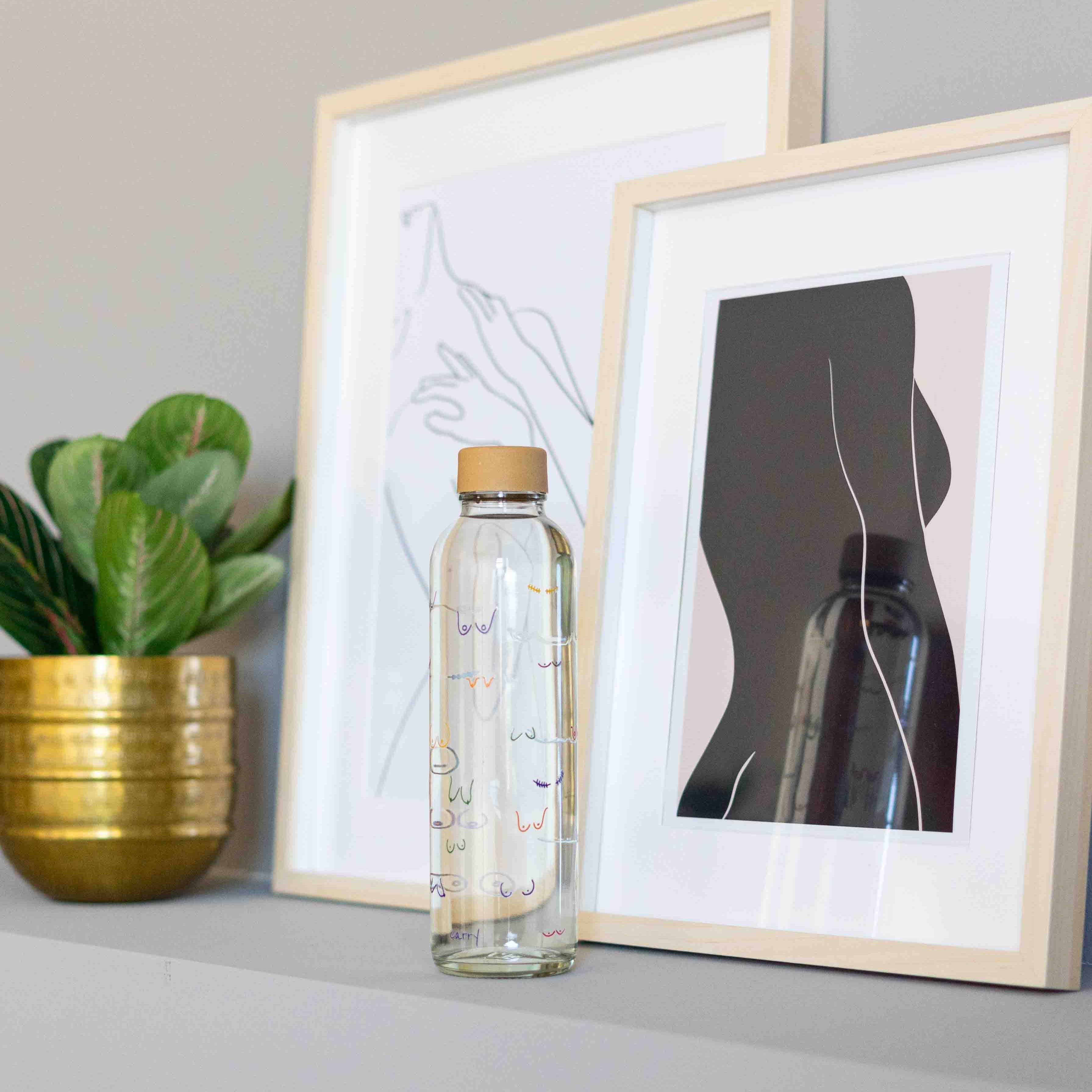 Eine Glasflasche steht vor zwei gerahmten Kunstwerken und einer Pflanze in einem goldenen Topf auf einem Regal.