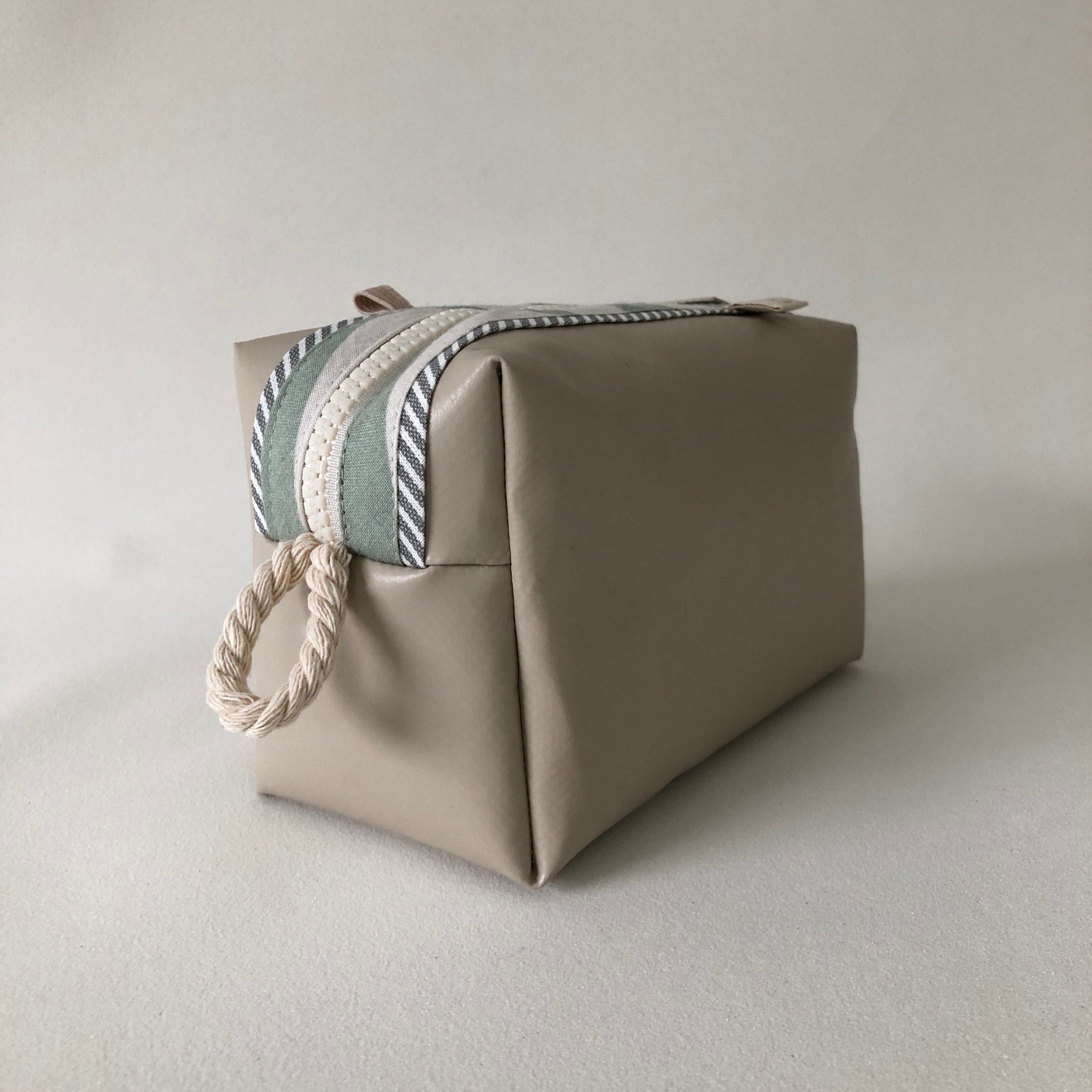Beige Kulturbeutel mit gestreiftem Reißverschluss und geflochtener Schlaufe auf einem hellen Hintergrund.