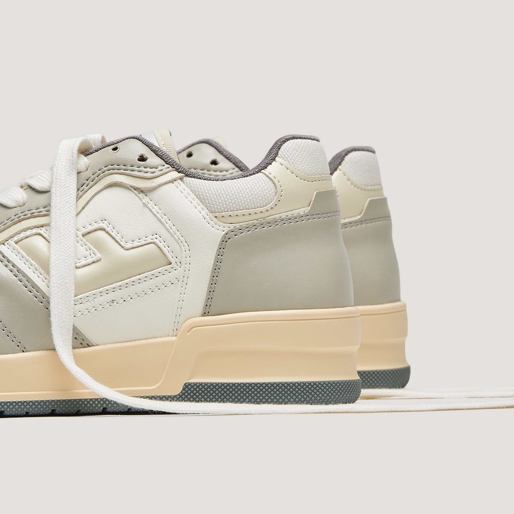 Beigefarbene Sneaker mit cremefarbenen Details und weißen Schnürsenkeln auf hellgrauem Hintergrund.