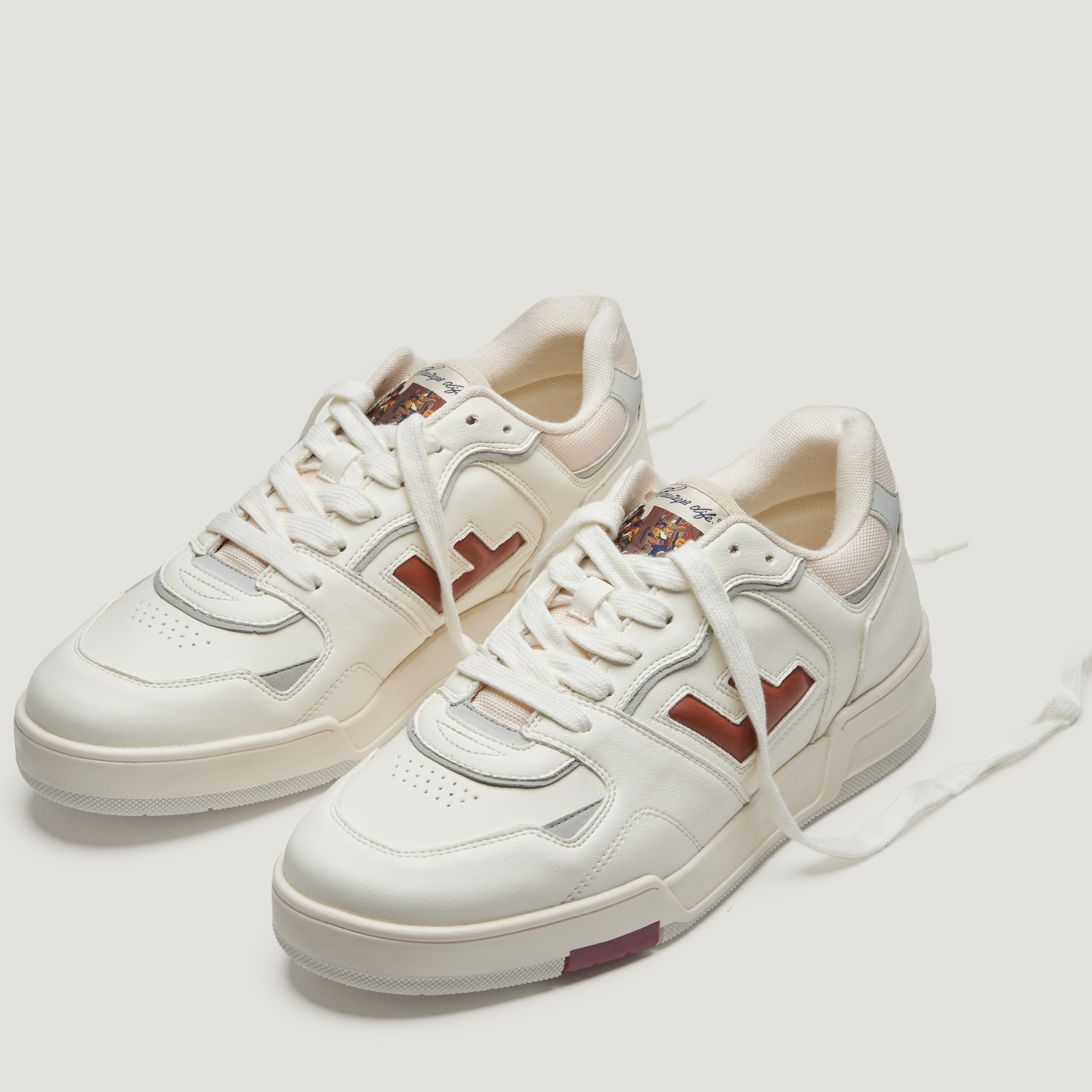 Weiße Sneaker mit braunen Details, weißen Schnürsenkeln und einem farbigen Logo auf der Zunge, auf hellgrauem Hintergrund.