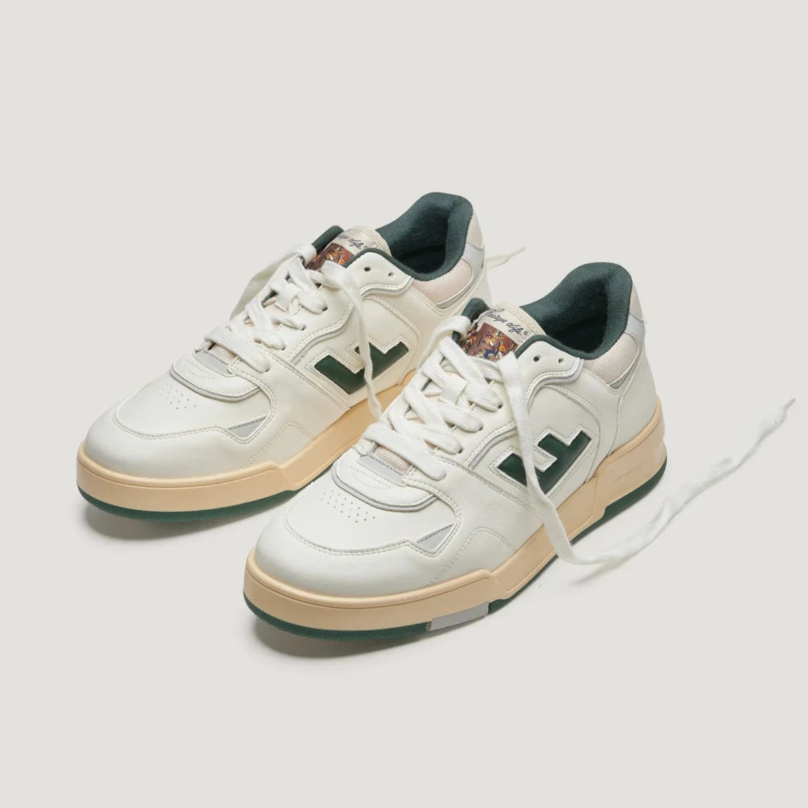 Weiße Turnschuhe mit beige und grünen Akzenten, schwarzen Logos und weißen Schnürsenkeln auf hellem Hintergrund.