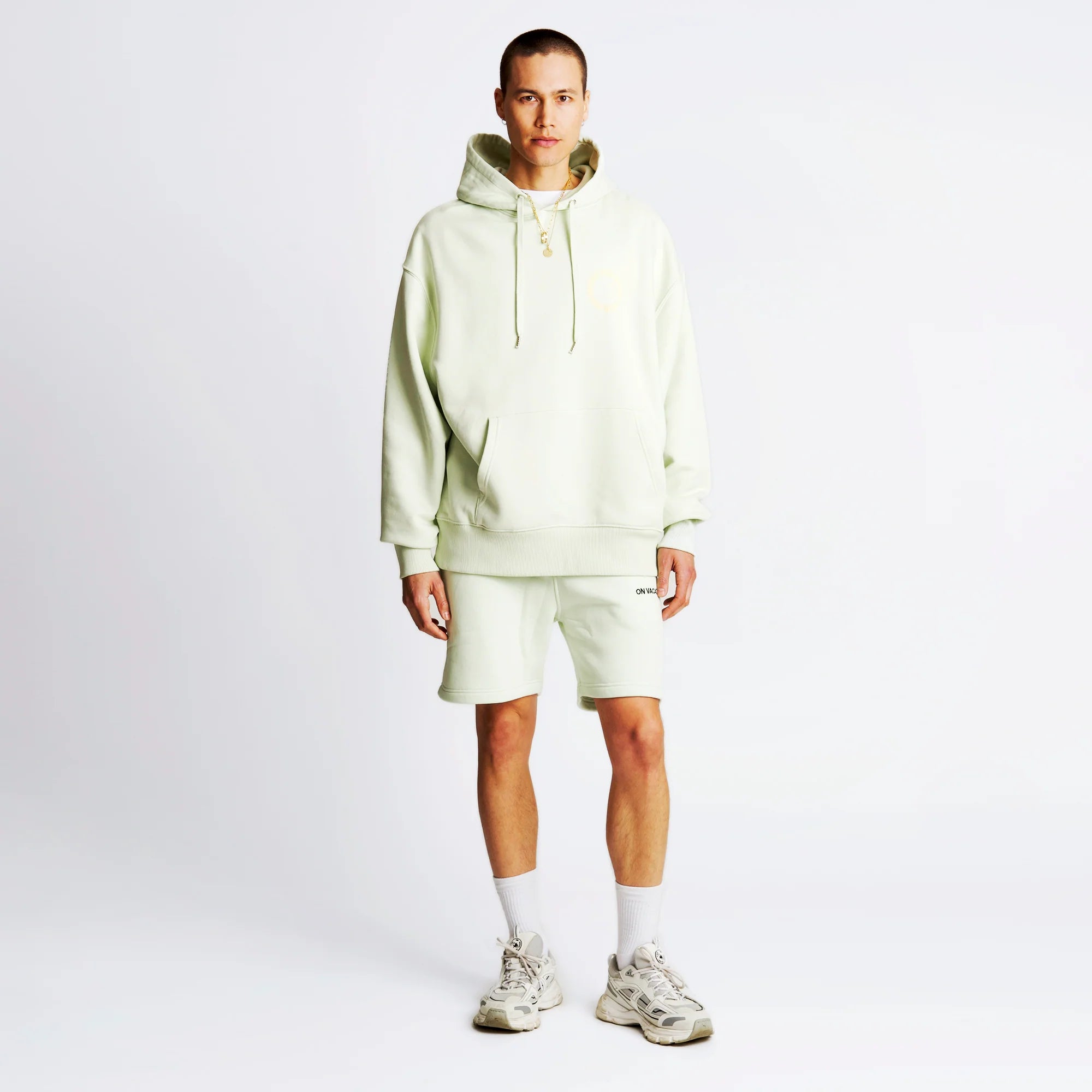 Person in cream-colored hoodie and shorts, with sneakers and socks, standing against a plain white background.
