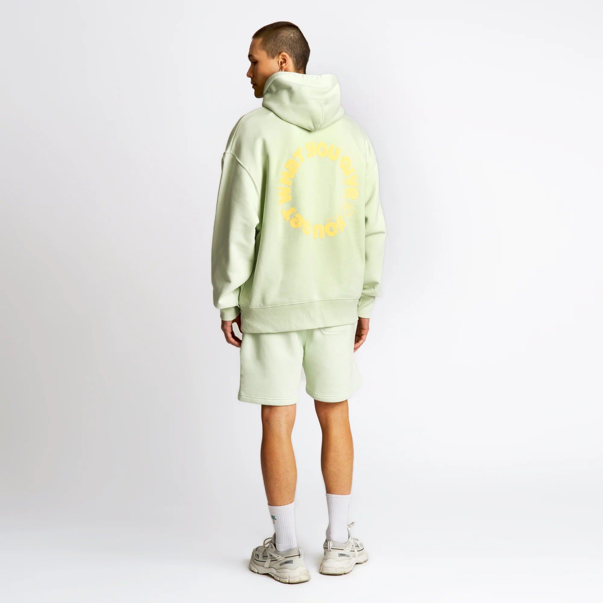 Mann im hellgrünen Hoodie und Shorts steht mit dem Rücken zur Kamera vor weißem Hintergrund.