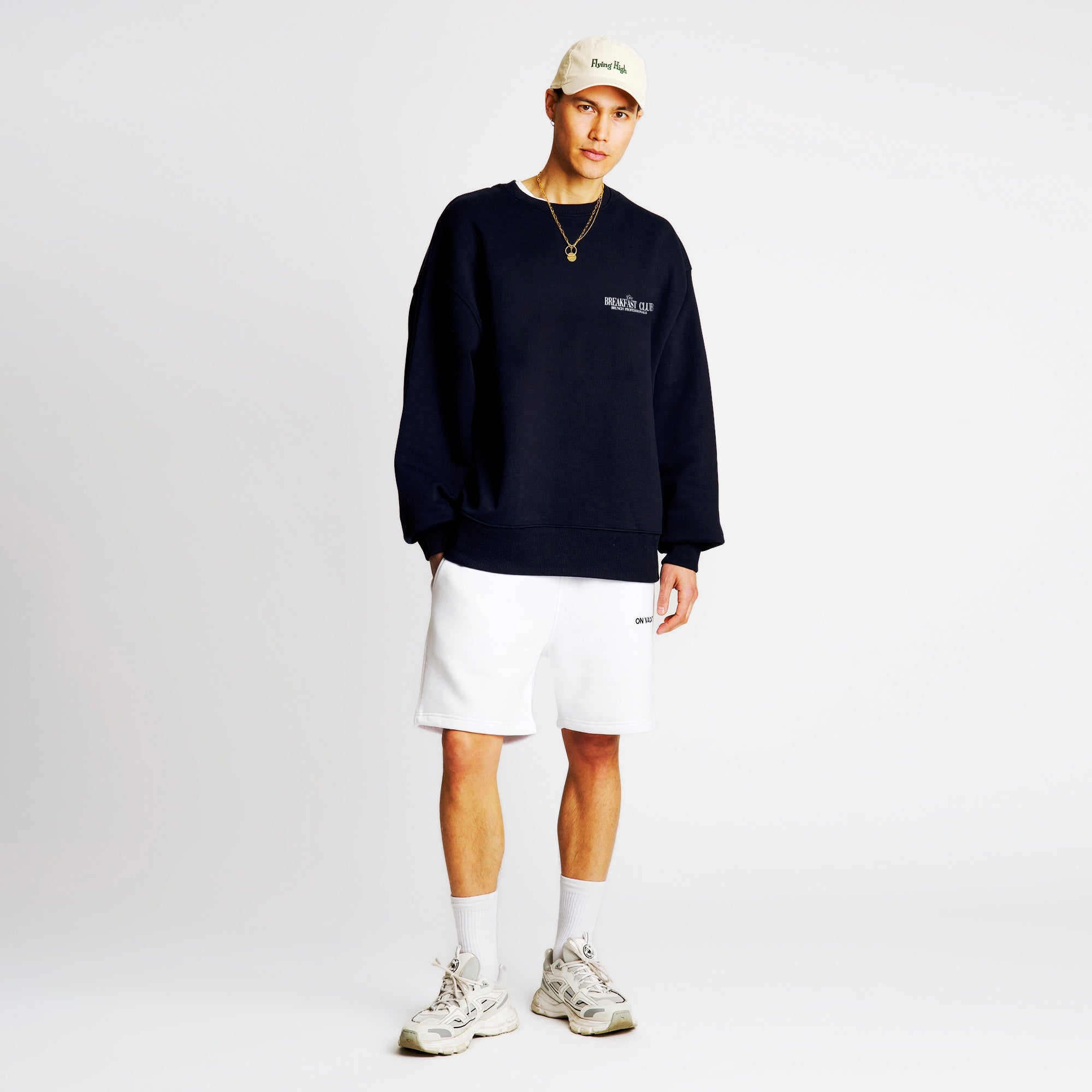 Ein junger Mann steht vor einem schlichten weißen Hintergrund und trägt eine beigefarbene Mütze, ein dunkelblaues Sweatshirt, weiße Shorts, weiße Socken und klobige weiße Turnschuhe mit einer goldenen Kette als Accessoire.