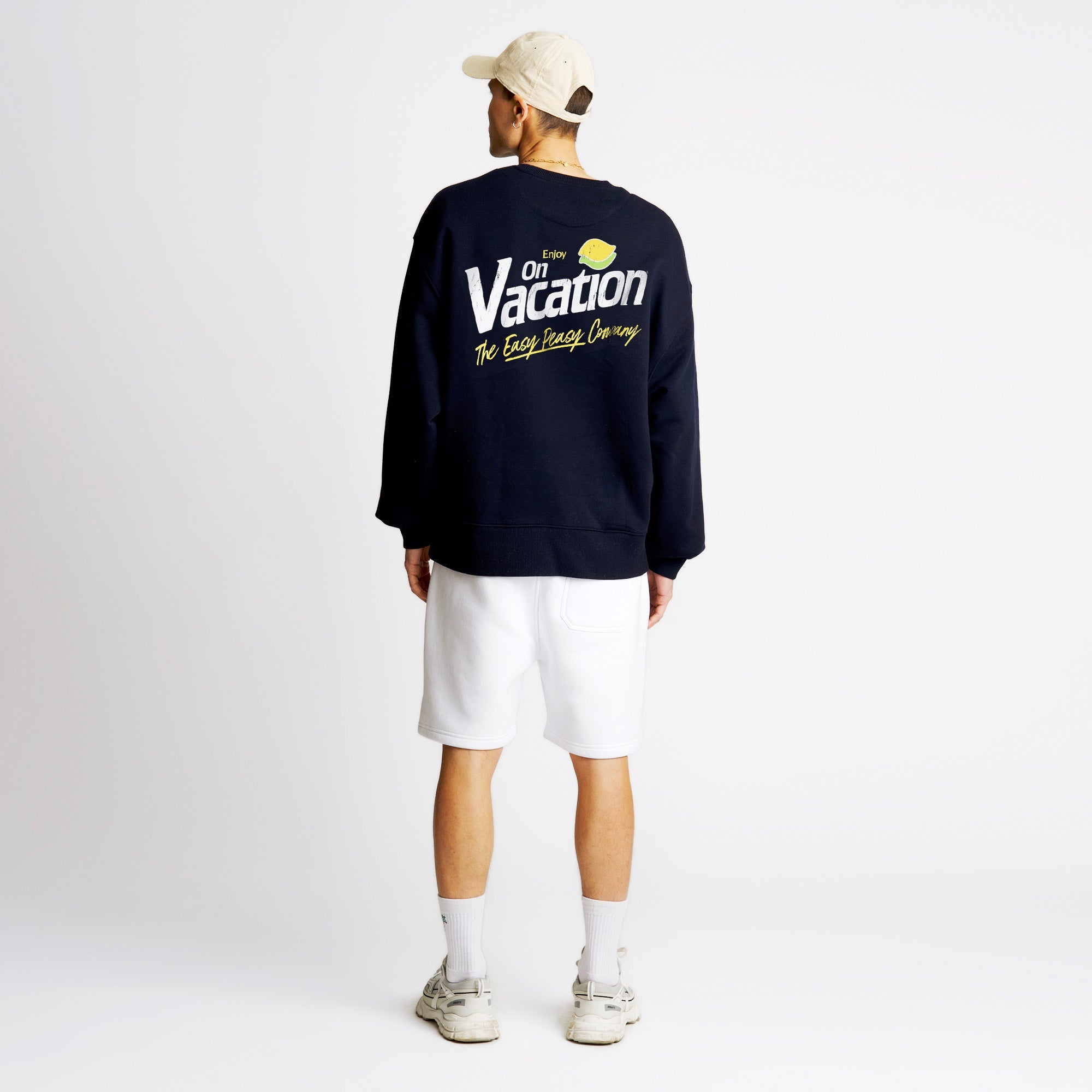 Eine Person, die eine beigefarbene Mütze, ein schwarzes Sweatshirt mit der Aufschrift On Vacation" auf dem Rücken, weiße Shorts und weiße Turnschuhe trägt, steht mit dem Gesicht nach außen vor einem schlichten weißen Hintergrund.