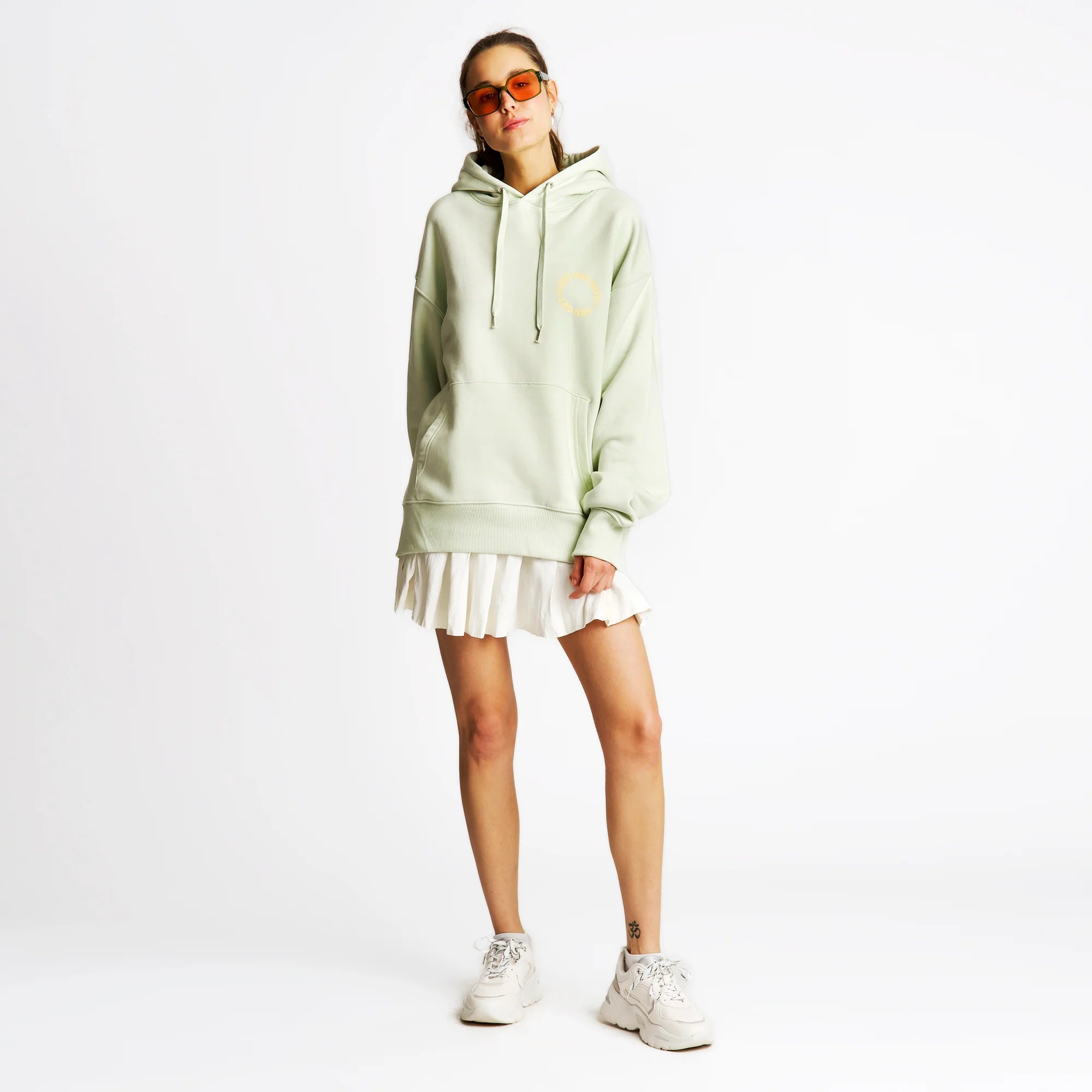Frau in hellem Kapuzenpullover, weißem Faltenrock und weißen Sneakers vor weißem Hintergrund.