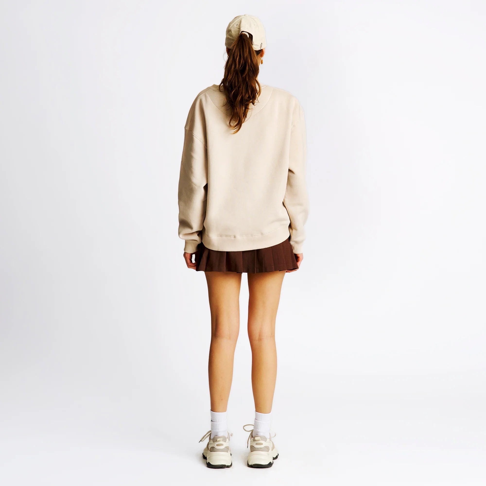 Frau von hinten in beige Pullover, braunem Rock, weißen Socken und Sneakers, trägt Mütze und hat langen Pferdeschwanz.