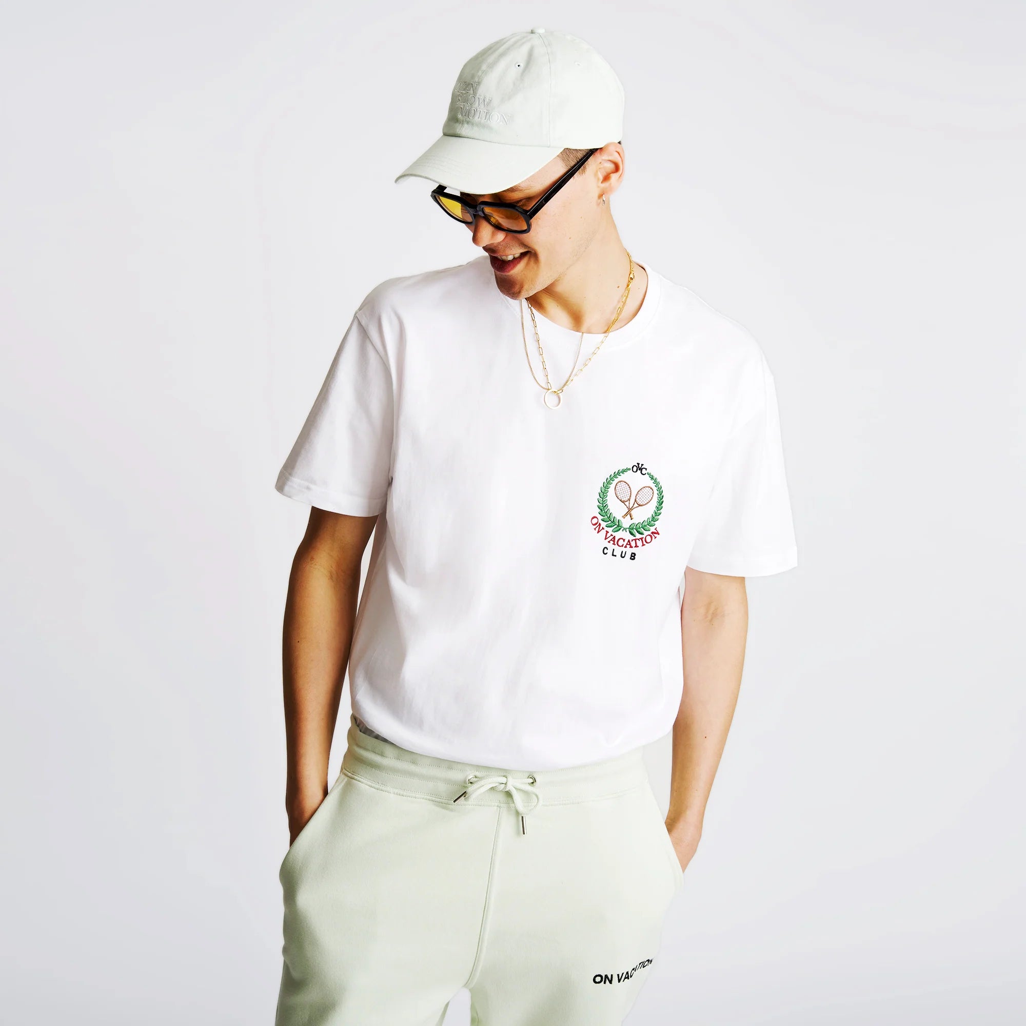 Person in weißem T-Shirt und heller Kappe schaut nach unten, trägt eine Halskette und eine Brille vor hellem Hintergrund.