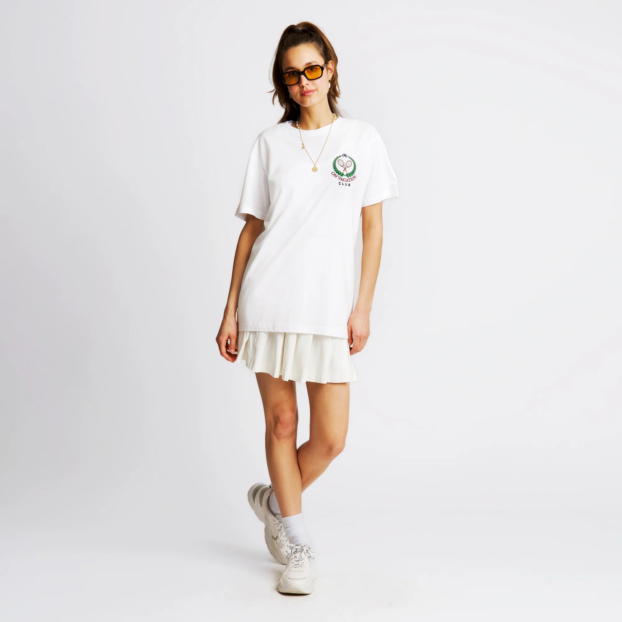 Frau in weißem T-Shirt und Rock, trägt Sonnenbrille und steht vor weißem Hintergrund.