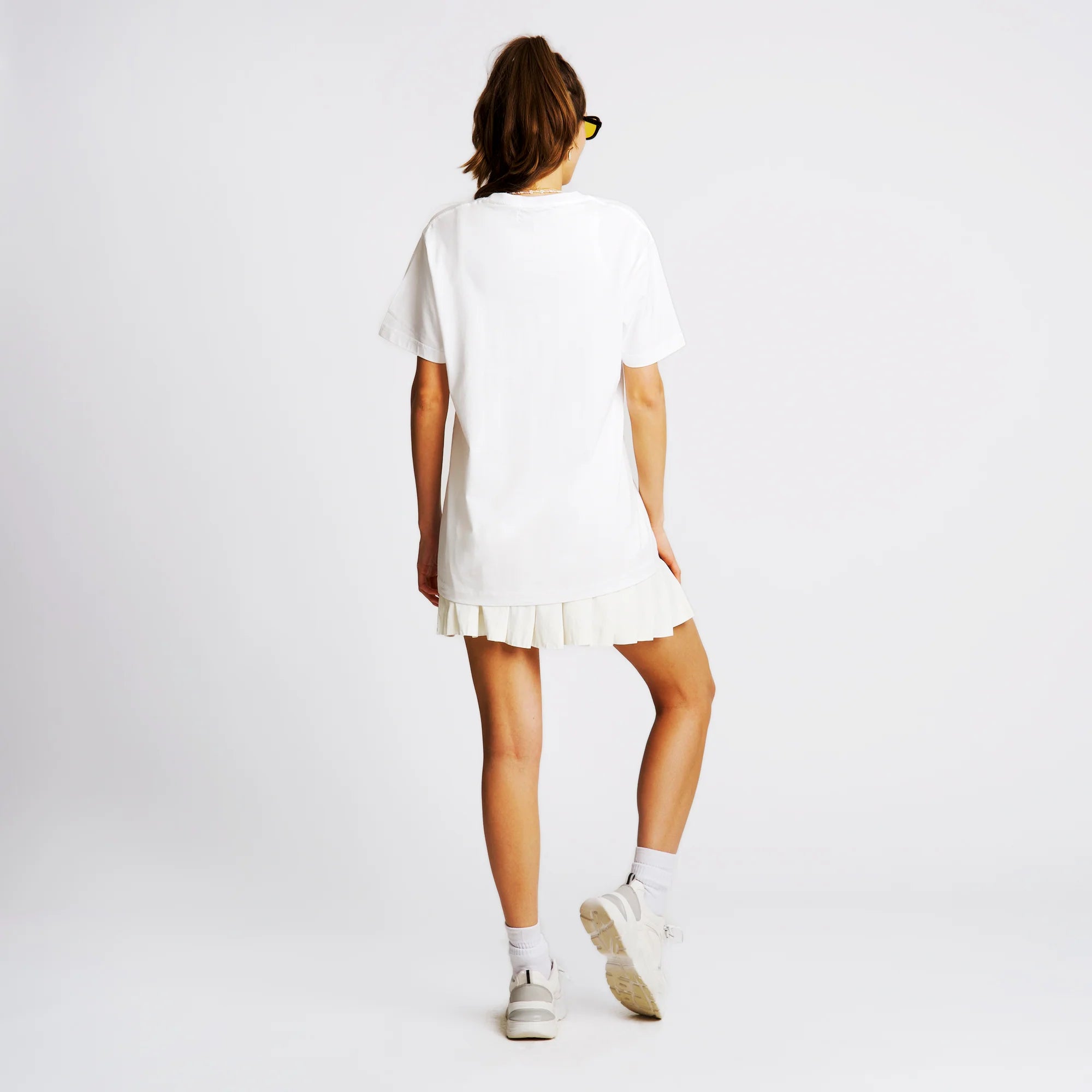 Person in weißem T-Shirt und Rock von hinten, trägt Sneakers und Socken auf weißem Hintergrund.
