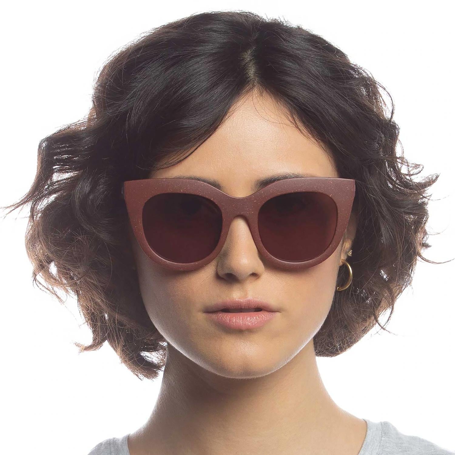 Eine Person mit kurzen, gewellten dunklen Haaren trägt eine große, runde, braune Sonnenbrille und einen kleinen goldenen Ohrring und schaut vor einem schlichten weißen Hintergrund direkt in die Kamera.