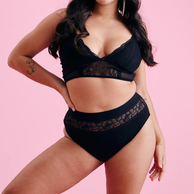 Eine Frau mit langen dunklen Haaren posiert selbstbewusst in schwarzen Spitzen-Dessous vor einem rosa Hintergrund. Sie hat eine Hand in die Hüfte gestemmt und einen neutralen Gesichtsausdruck, der das Dessous-Set zur Schau stellt.