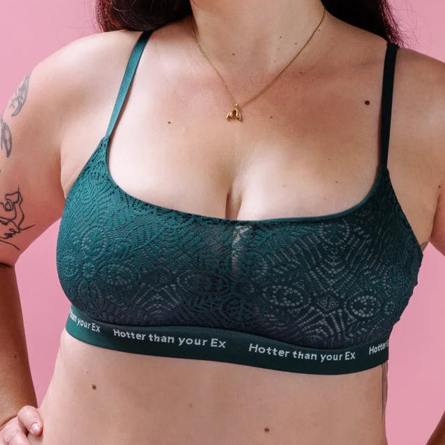 Eine Frau, die einen grünen Spitzen-Bralette mit dünnen Trägern trägt, steht vor einem rosa Hintergrund. Auf dem Band des Bralettes steht in weißen Buchstaben Hotter than your Ex. Sie trägt eine Halskette und sichtbare Tattoos auf ihrem Arm.