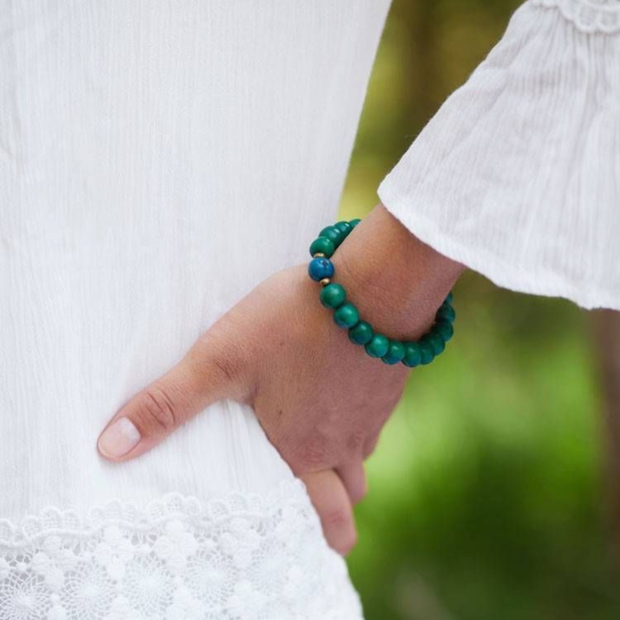 Eine Person, die ein weißes, langärmeliges Kleid mit Spitzenbesatz trägt, stützt ihre Hand auf ihre Hüfte und zeigt ein grünes Perlenarmband an ihrem Handgelenk. Der Hintergrund ist unscharf mit grünen Andeutungen.
