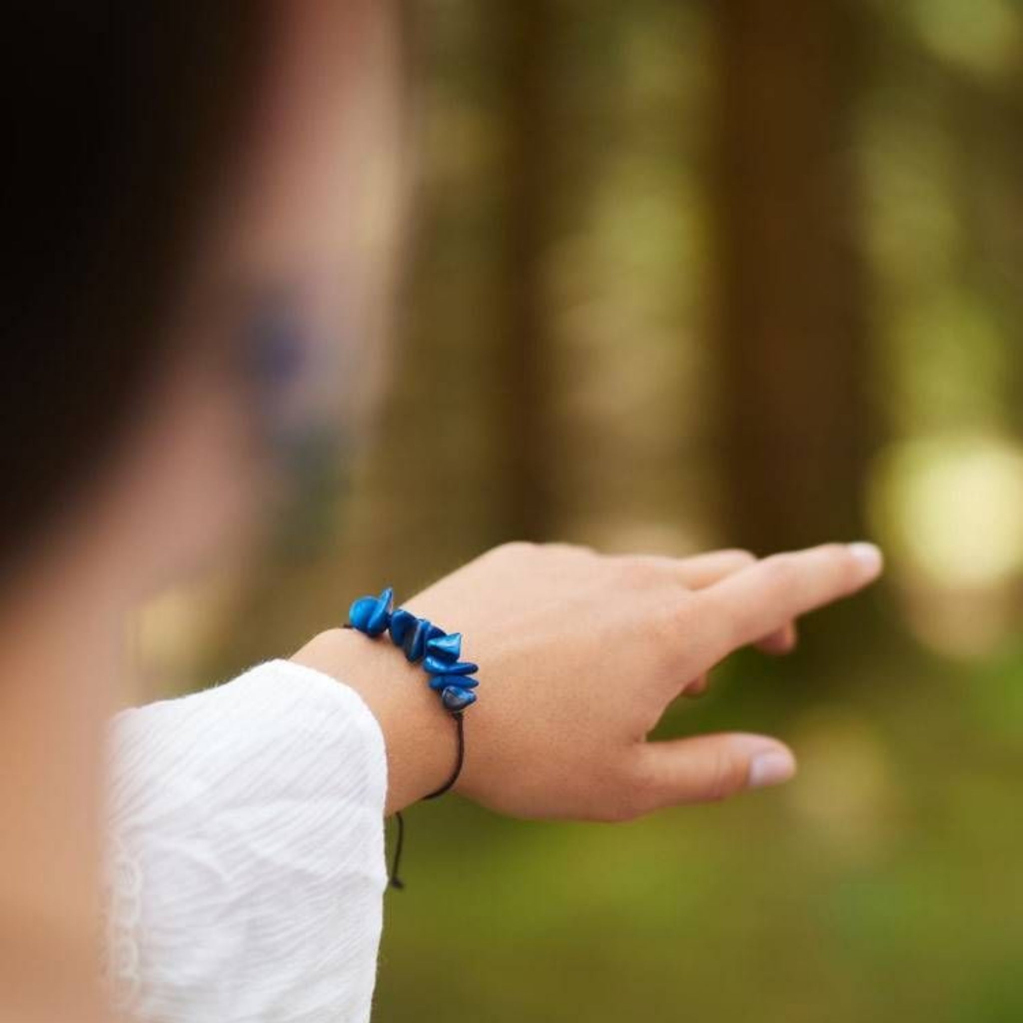 Eine Person, die einen weißen Ärmel und ein Armband mit blauen Steinen oder Perlen trägt, zeigt auf einen unscharfen, grünen Waldhintergrund. Der Fokus liegt auf ihrer ausgestreckten Hand und ihrem Armband.