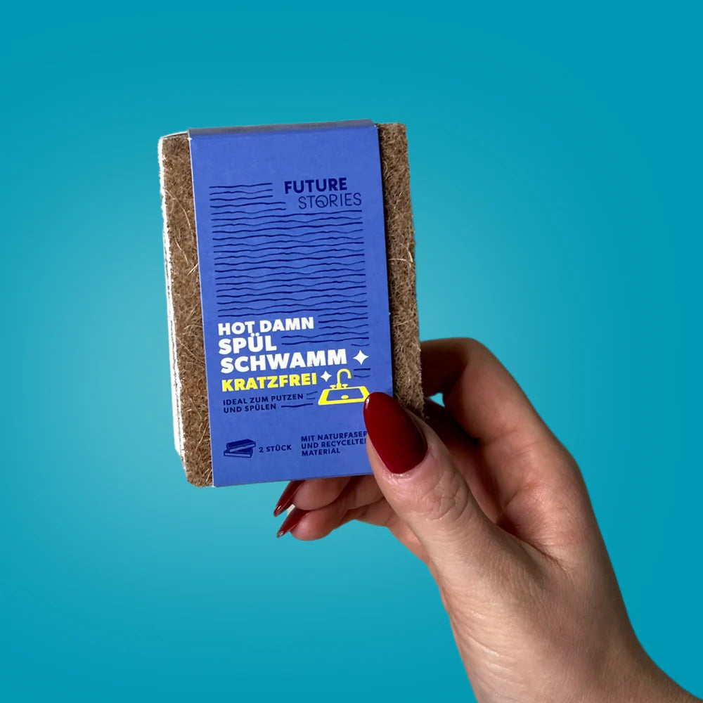 Eine Hand mit rotem Nagellack hält einen braunen Küchenschwamm in einer blauen Verpackung mit der Aufschrift "Future Stories Hot Damn Spülschwamm Kratzfrei!" vor einem einfarbigen tealfarbenen Hintergrund.