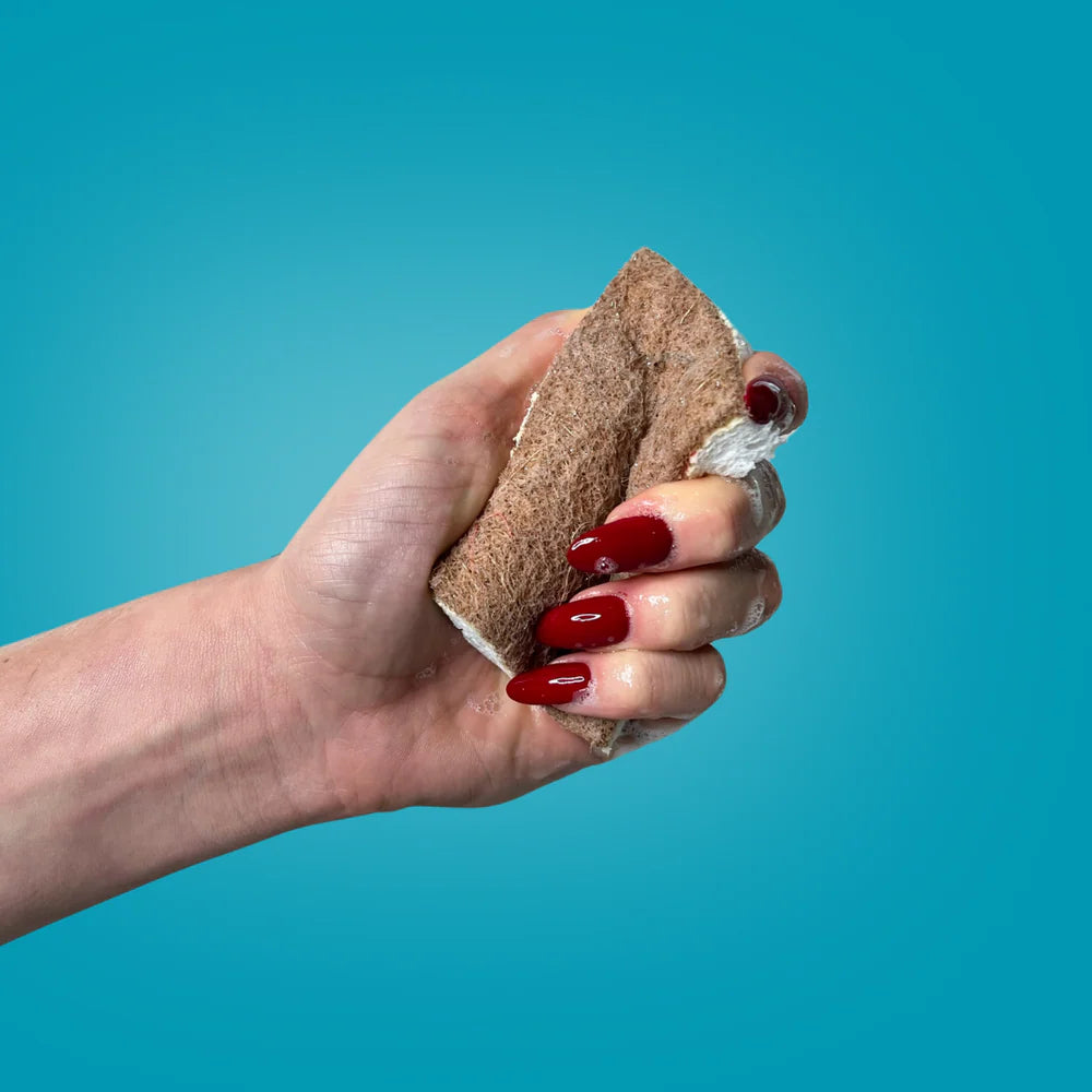 Eine Hand mit langen roten Nägeln hält einen braunen Naturschwamm mit Seifenlauge vor einem schlichten blauen Hintergrund.