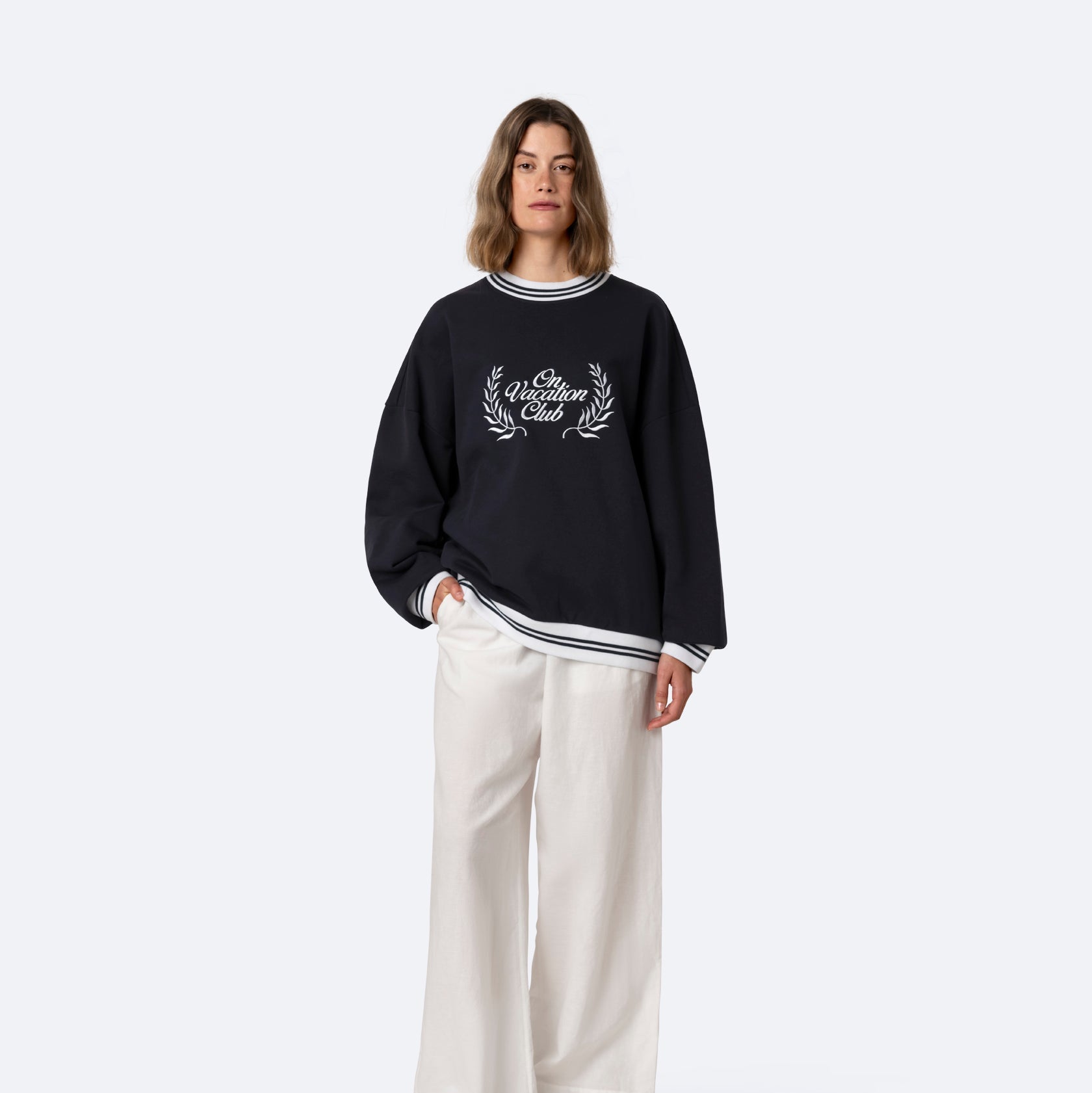 Eine Person steht vor einem schlichten Hintergrund und trägt einen lockeren schwarzen Pullover mit dem Text "Club William" und einem Lorbeermuster, eine weiße Hose mit weitem Bein und beigefarbene Slipper.